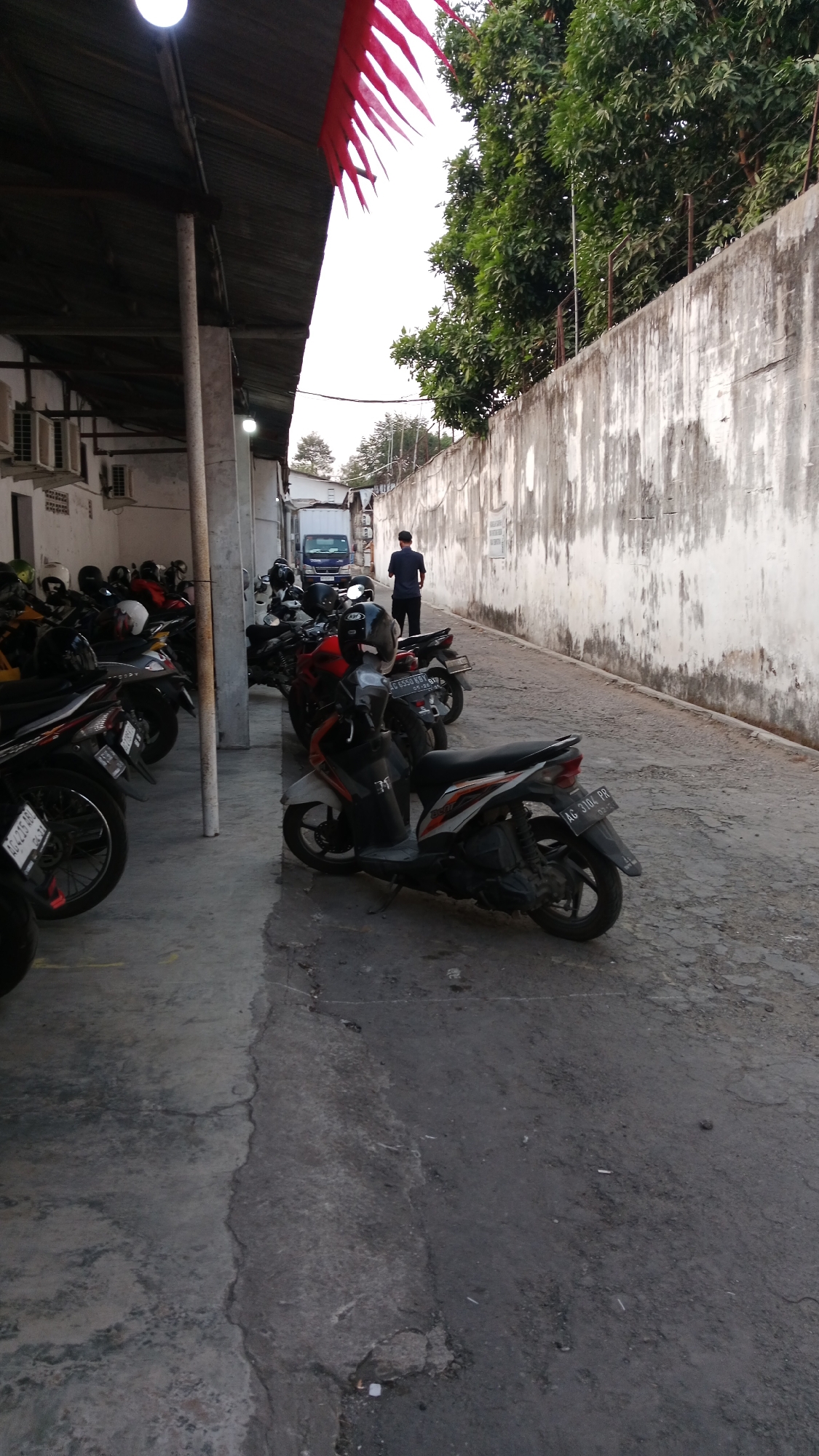 The image depicts a parking area located in Blitar, Indonesia. The scene shows a collection of parked motorcycles and a blue truck in the background. A person in a dark blue shirt and black pants is standing near the truck. The parking area is paved with concrete and asphalt, with a large concrete wall on the right side of the frame. Red flags are visible hanging from a covered structure on the left. The environment appears to be outdoors, with trees visible behind the wall.