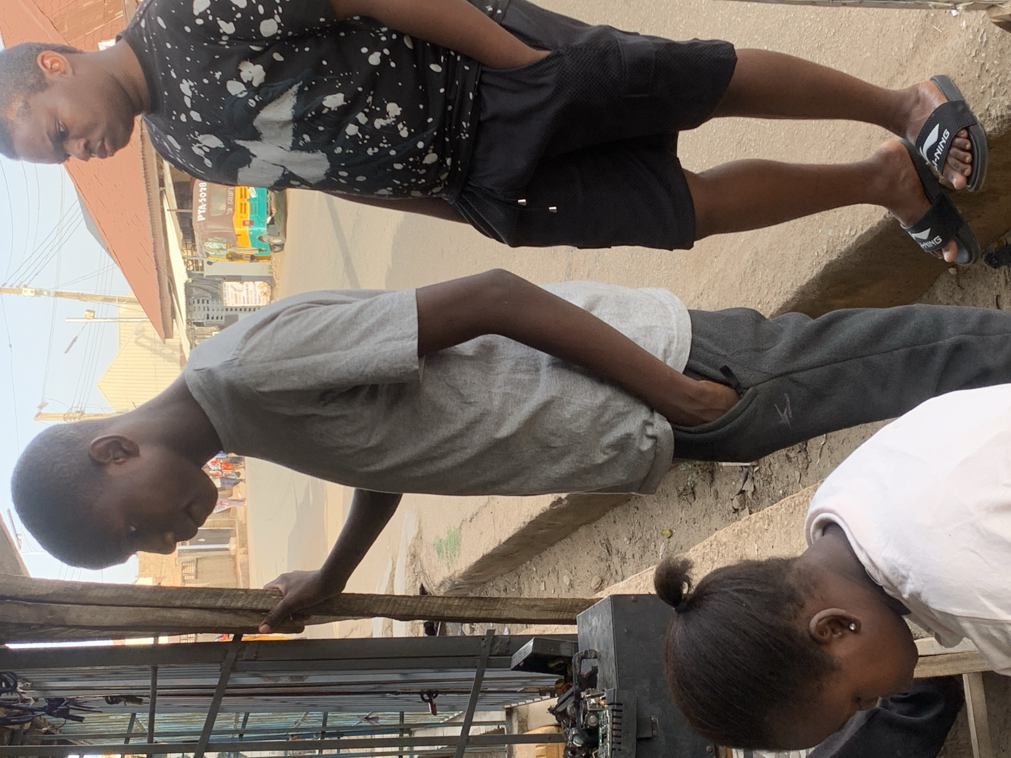 The image depicts three young individuals on what appears to be a street in Jos, Nigeria. One person is wearing a black shirt with white splatter design and black shorts, and stands on the right side of the frame. A second person is wearing a grey t-shirt and grey sweatpants and leans over something, seemingly interacting with it. A third person is wearing a white shirt and has a ponytail. Behind them is a metal structure, possibly a storefront or stall. The environment is an urban setting, with buildings and a street visible in the background.