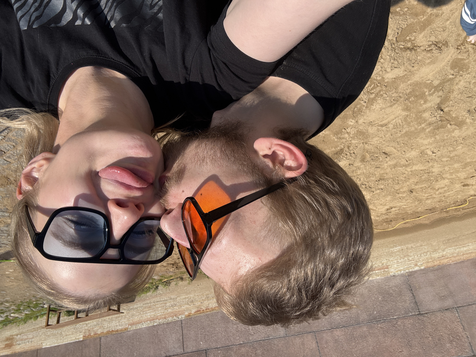 Two individuals are lying next to each other in an outdoor setting. One person has blond hair, is wearing black sunglasses and has their tongue sticking out. The other has short brown hair, a beard, and orange-tinted sunglasses. They are wearing black shirts. They are lying on a surface of sand, with an edge of brick paving visible on the right and bottom. The location is likely Saint Petersburg, Russia, based on the architectural style of the background elements and materials like brick pavers.