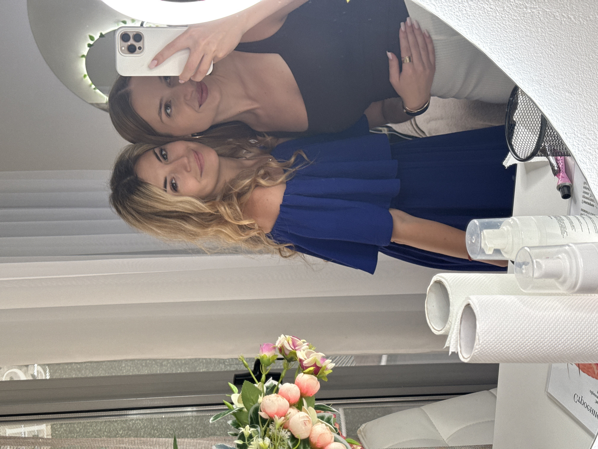 Two women are taking a selfie in a mirror. One woman has blonde hair and is wearing a blue dress. The other woman has brown hair and is wearing a black top. The mirror is surrounded by a white frame and a half-moon shaped light. In front of the mirror are flowers, white paper rolls, and cosmetic bottles. A window with blinds is visible in the background. The Cyrillic text on a card suggests the location is likely in Budva, Montenegro.