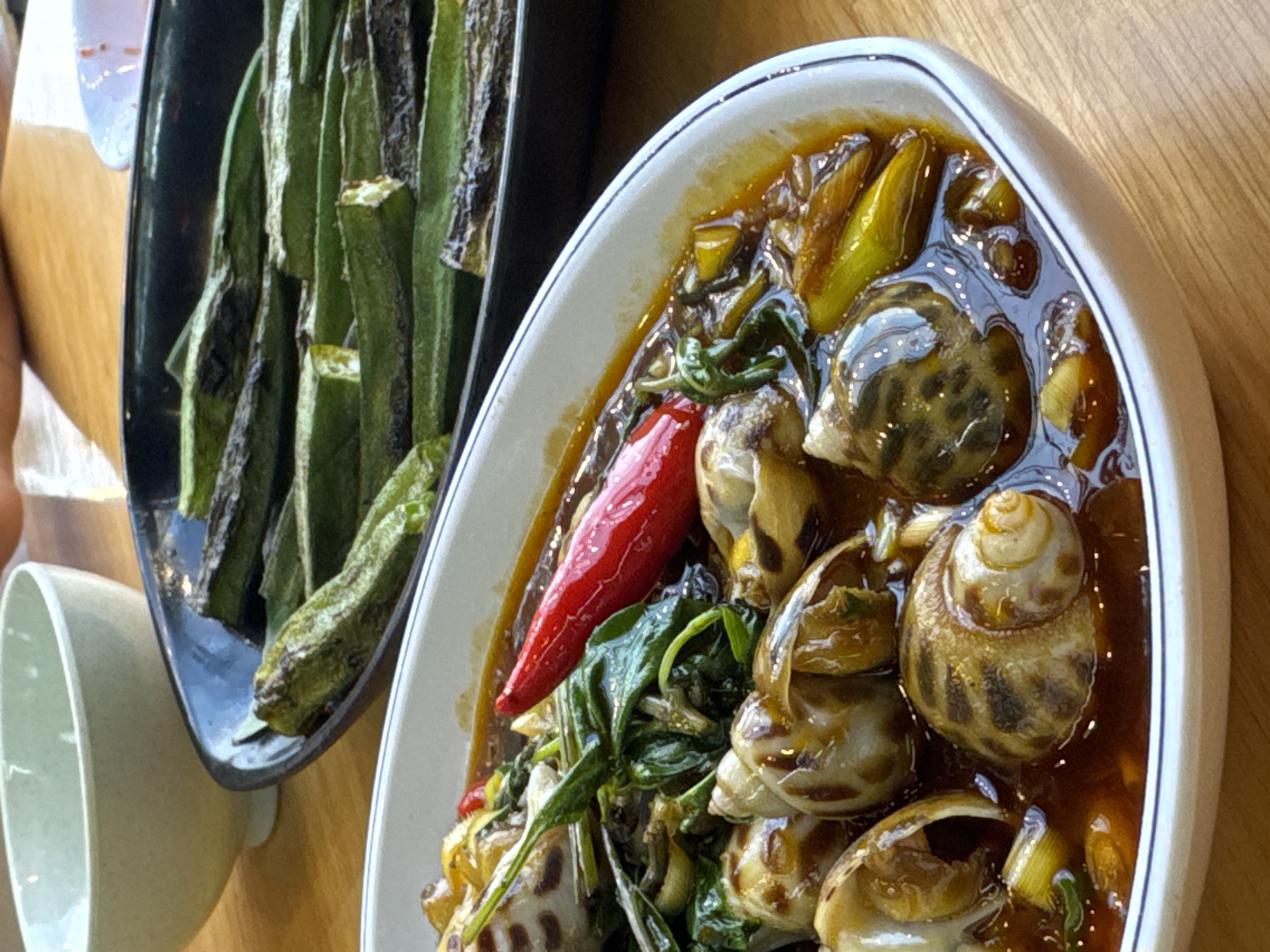 A close-up shot reveals a dish of snails swimming in a dark sauce, garnished with a red chili pepper and green herbs, on a white oval plate next to a black dish of sautéed okra. The plates rest on a wooden surface, and a white bowl is visible in the lower left corner. The photo was taken at a restaurant in Đà Nẵng, Vietnam.