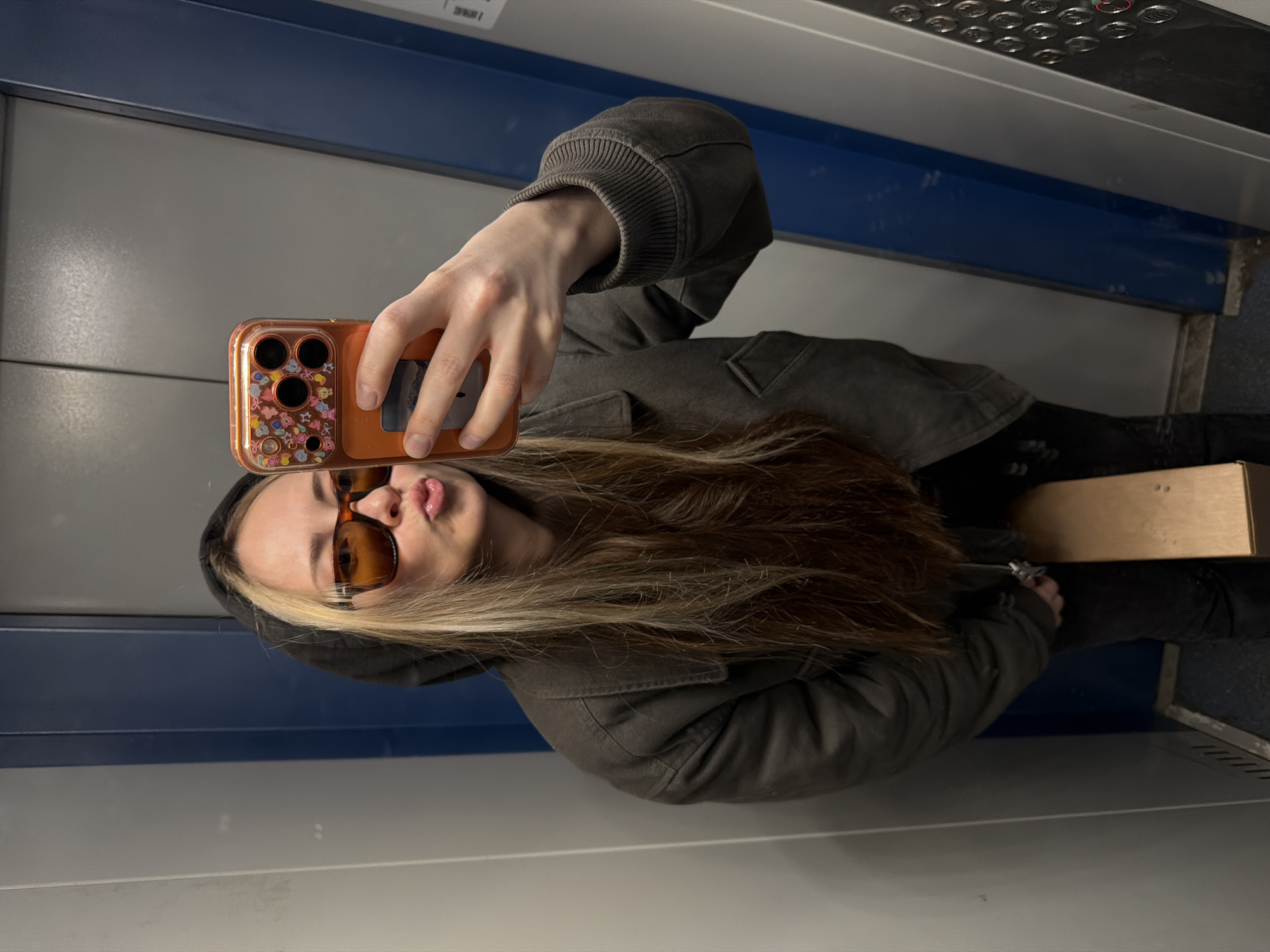 A female taking a selfie in an elevator. She has blonde hair and is wearing sunglasses, a hoodie and a jacket. She is holding an orange phone with a sticker-covered case. The elevator has blue and grey panels with a button panel at the top. The location is unknown; no details in the image indicate it is Khimki, Russia.