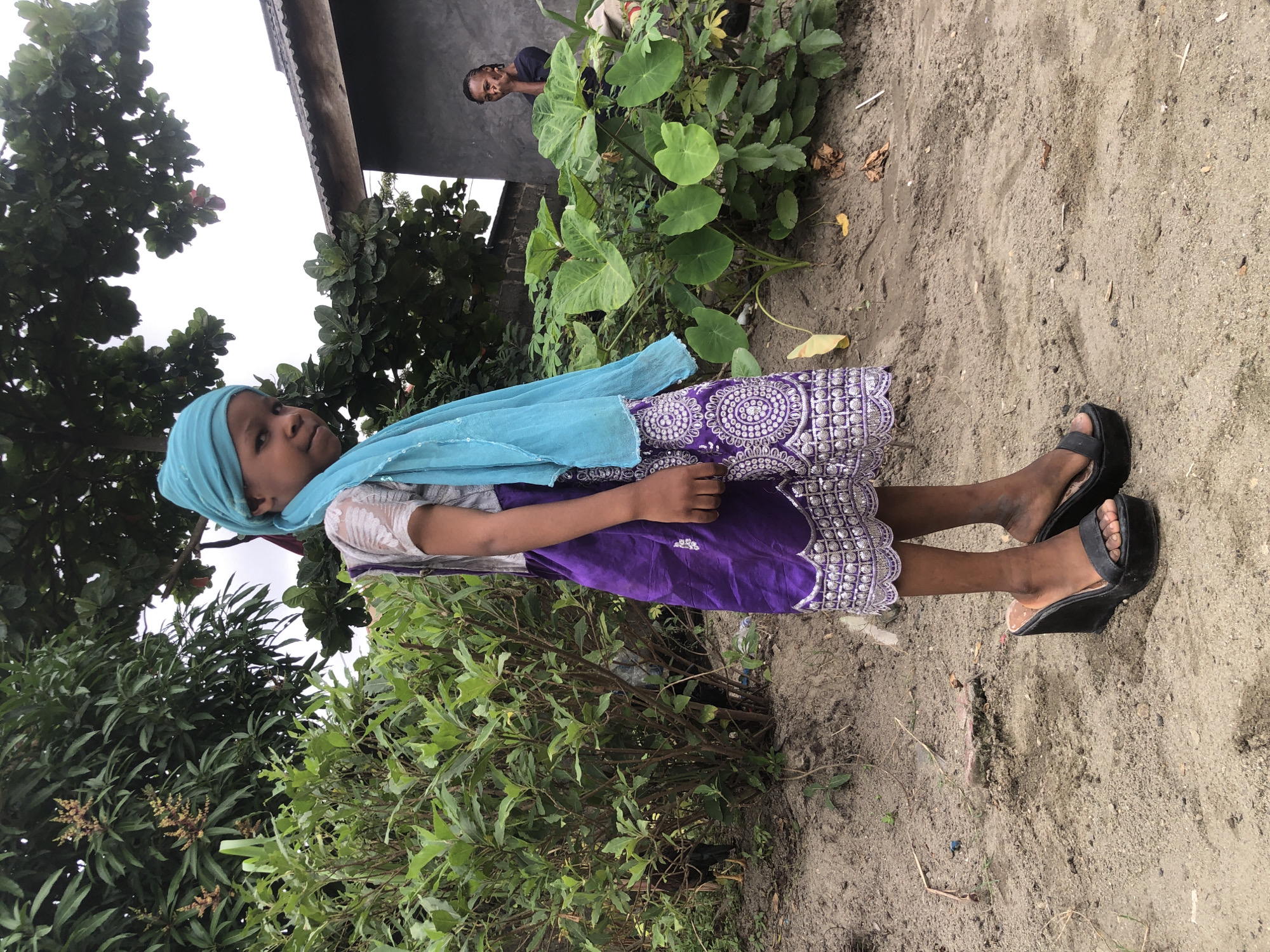 In Ado, Nigeria, a young girl stands in a sandy area, adorned in a bright blue headscarf and a purple dress with intricate silver patterns. Her dress features a wide, ornate border at the hem. She is wearing black wedge sandals. To her left, lush green bushes and trees form a dense backdrop, with some foliage showing yellowing leaves. In the background, a simple, grey building with a corrugated metal roof is visible, with another person, an adult male, standing near it, looking towards the camera and making a gesture with his hand. The ground is primarily composed of sand with scattered dry leaves and small stones. The sky above is overcast.