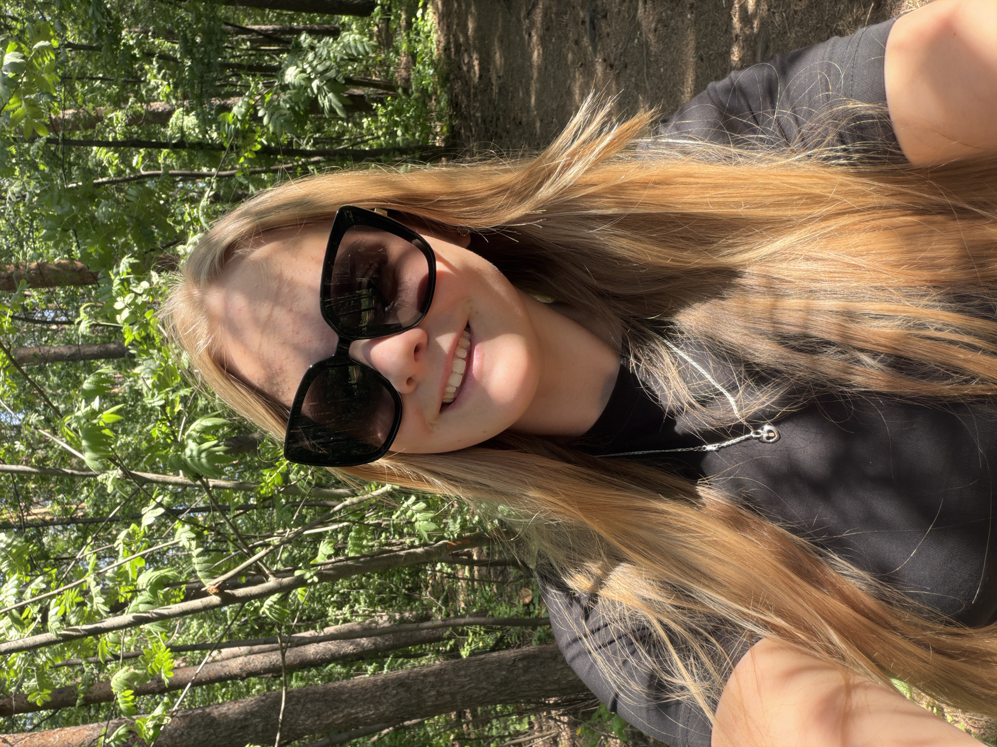 A young woman with long, blonde hair is smiling in a selfie. She is wearing sunglasses and a black shirt, with a silver necklace visible. The background shows a wooded area with trees and sunlight filtering through the leaves. The location is outside, possibly in a park or forest setting, but no specific identifiable elements suggest Izhevsk, Russia.
