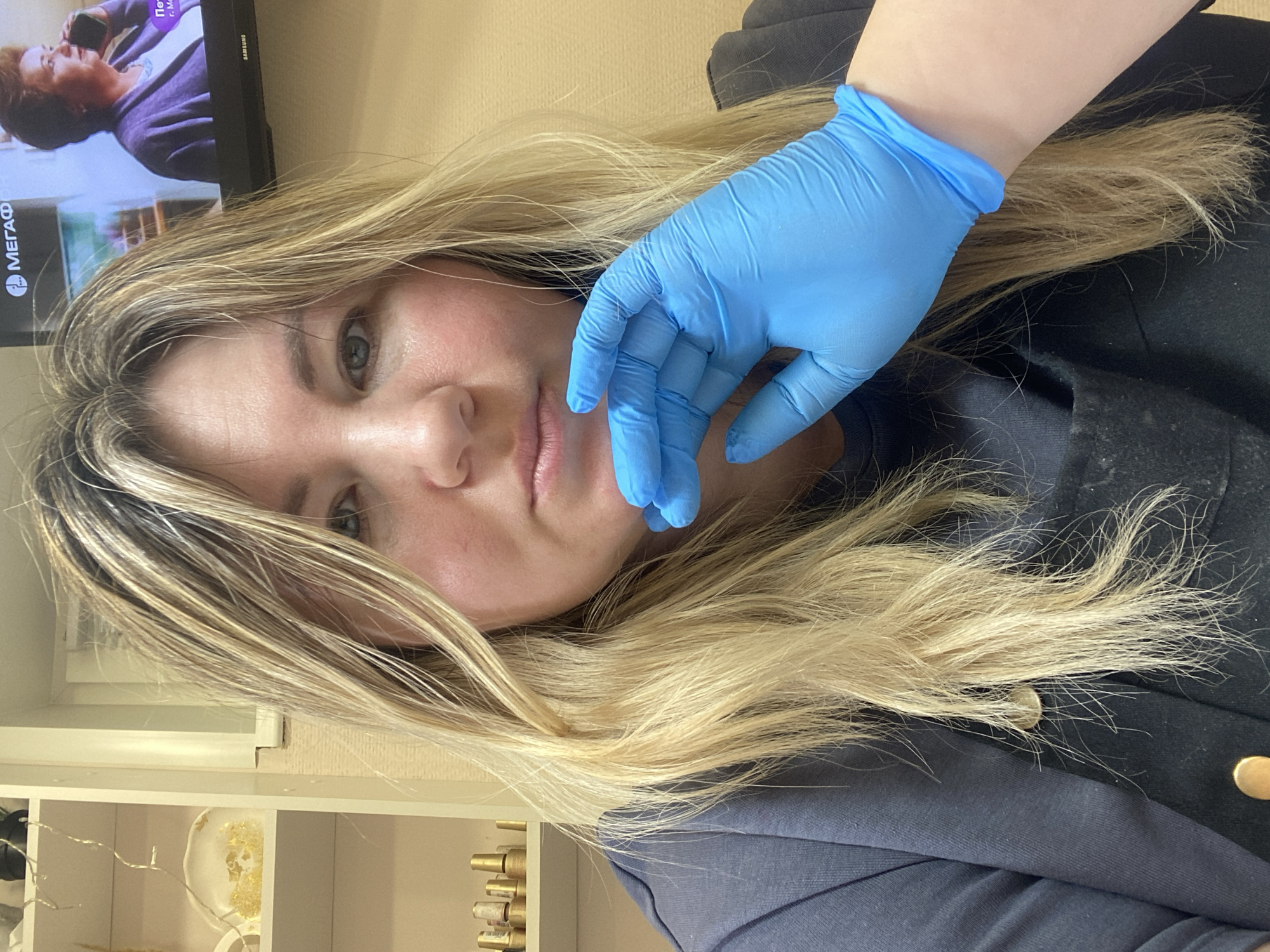 The image captures a close-up, slightly tilted portrait of a woman with long, wavy blonde hair, looking directly at the viewer with a neutral expression. Her left eye, which appears blue or greenish-gray, is visible, along with parts of her nose and mouth. A hand wearing a bright blue medical-style glove is prominently placed in front of her face, resting near her cheek and lips, suggesting a treatment or procedure.
The woman is wearing dark attire, including what appears to be a dark jacket or top with a gold button visible on the shoulder, and a dark blue or gray garment underneath. Her blonde hair shows various shades, possibly indicating highlights or a natural blend of colors, and appears to be somewhat styled or tousled.
In the background, a television screen displays a program featuring a woman with dark hair in a purple top, seemingly engaged in a phone conversation. Visible text on the screen includes "МЕГАФОН" (MEGAFON), "г. М..." (likely "city M..."), and "Пст...". The "Samsung" brand name is visible on the television's frame. Below the TV, to the left, a white shelving unit with various items, including gold-colored decorative pieces and string lights, is partially visible. The indoor setting, along with the gloved hand, suggests an environment such as a beauty salon, clinic, or a dedicated home treatment area.