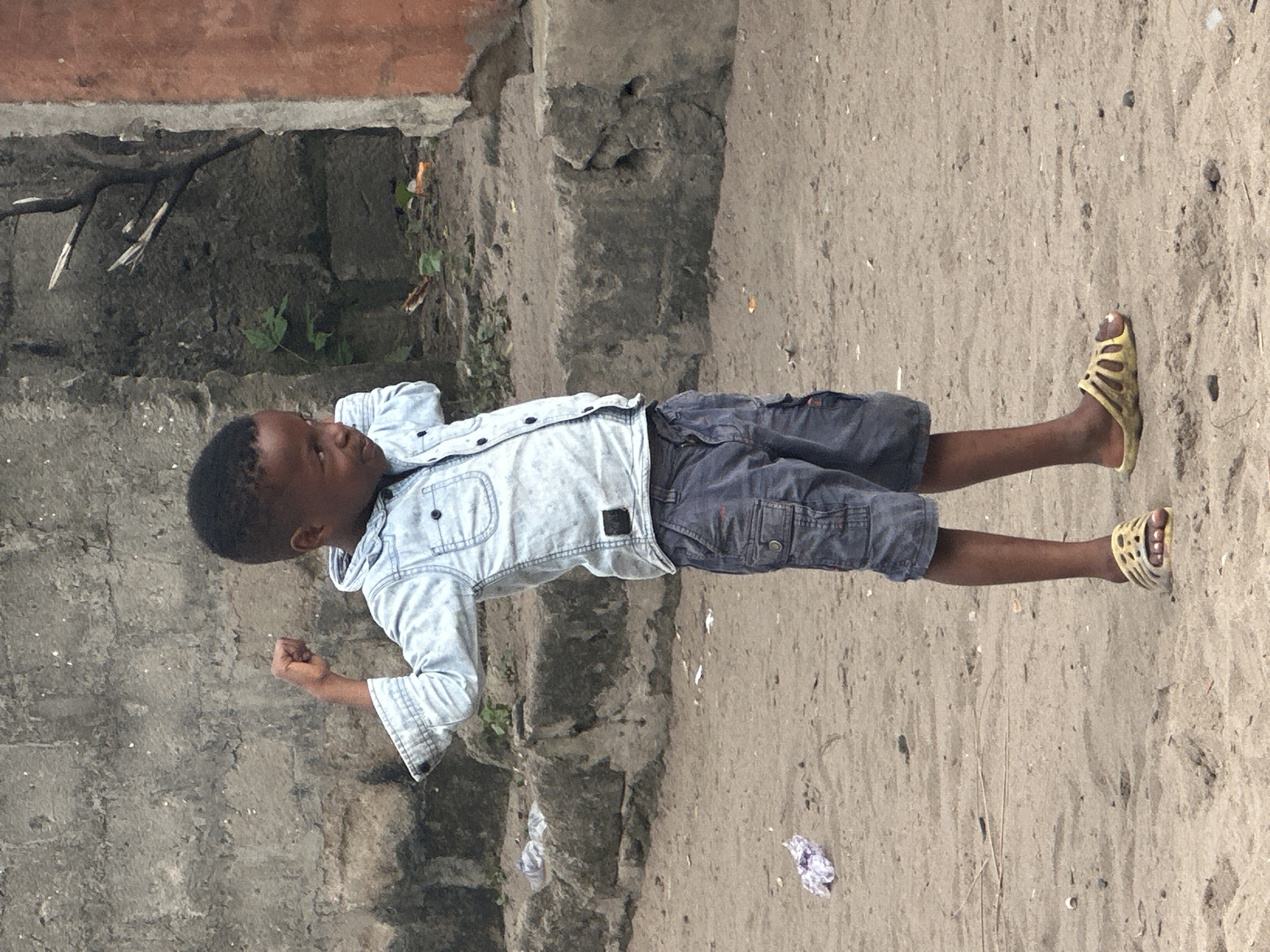 A young Black boy stands in a dirt area in Badagry, Nigeria, wearing a light blue denim shirt and grey cargo shorts. His left arm is bent at the elbow, with his fist clenched and held outwards, and his head is turned to his left, looking slightly down. The background consists of a rough concrete wall and more sandy ground. There is some scattered debris on the ground, including a crumpled piece of purple and white material to the boy's left and some twigs and leaves. The boy is wearing yellow slide sandals.