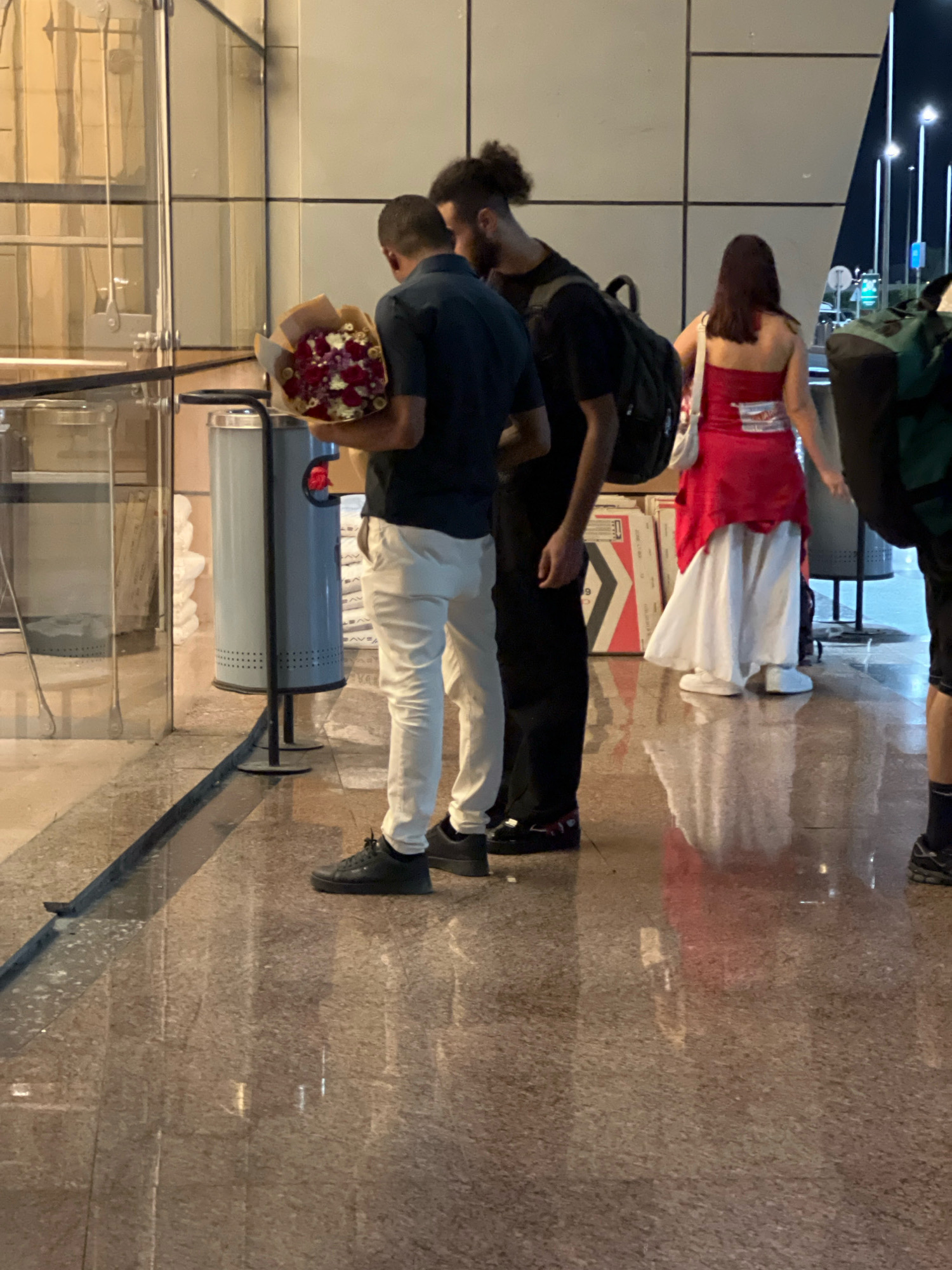 In a city setting resembling Sharm El Sheikh, Egypt, a group of people stand on a polished, reflective floor. In the foreground, a person in a dark shirt and white pants holds a bouquet of red and white flowers. To their right, another person, wearing a dark t-shirt and black pants, carries a black backpack. Further to the right, a person in a red strapless top and a long white skirt walks away from the camera, carrying a white bag over their shoulder. In the background, a cylindrical gray trash can with perforated sides is visible. The environment appears to be an indoor public space, possibly an airport or a hotel lobby, with large glass panels and textured walls. Lights from street lamps are reflected on the floor.