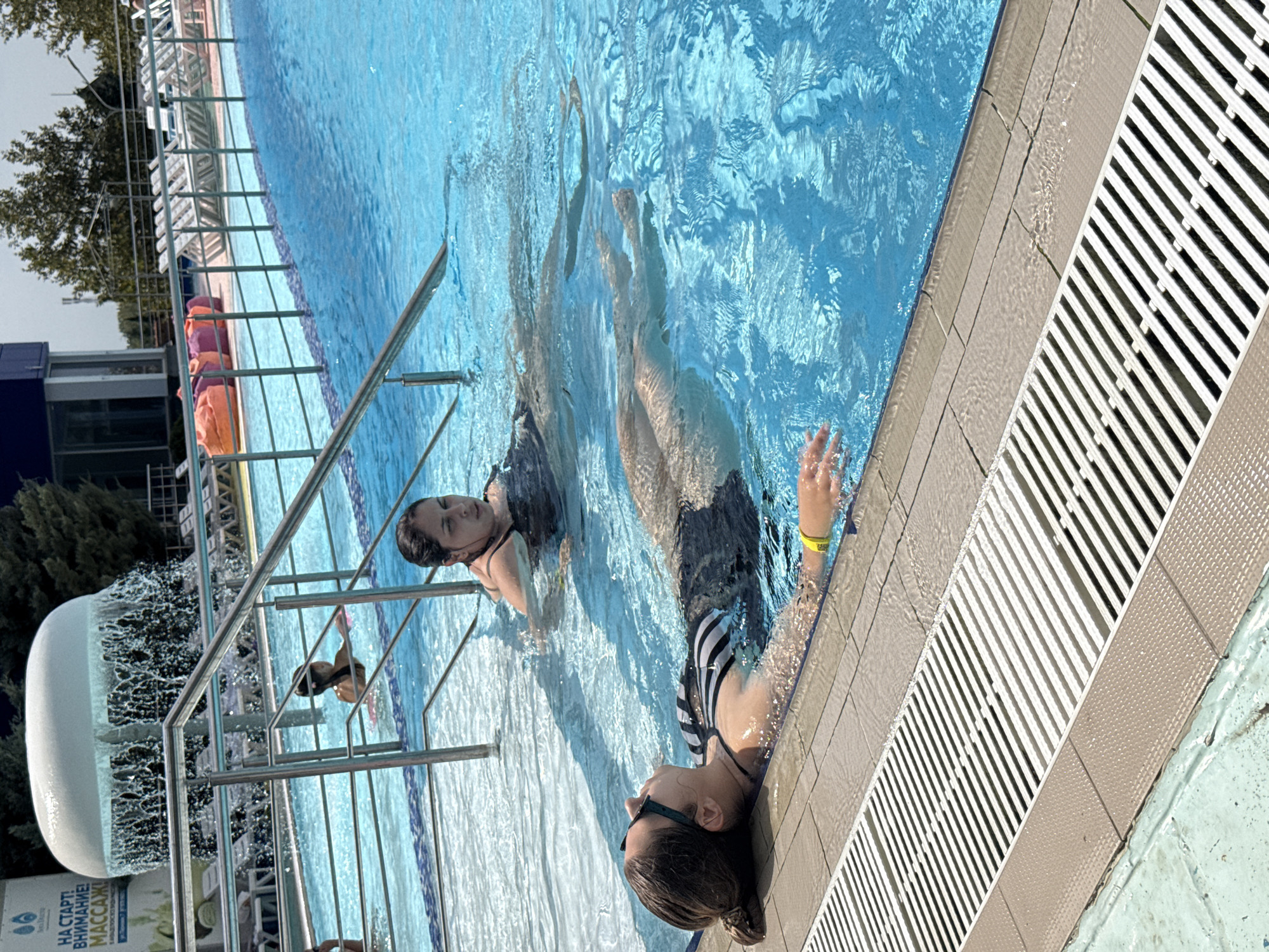 A woman is floating on her back in a swimming pool. She is wearing a black and white striped swimsuit and sunglasses. Her arms are extended above her head. Another woman is in the background, swimming in the pool. The pool is blue and has a tiled edge. In the background, there are lounge chairs and trees. The image appears to have been taken on a sunny day.