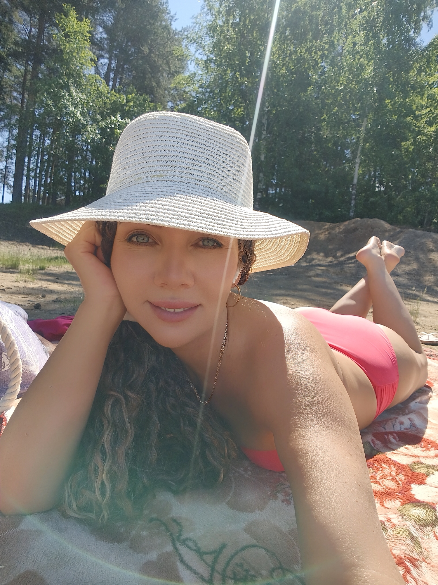 A woman wearing a wide-brimmed straw hat and a pink bikini is lying on her stomach on a blanket on a sandy beach. She is resting her head on her hand and looking towards the camera with a smile. The background shows trees and a clear blue sky. The scene appears to be outdoors, possibly during a sunny day.