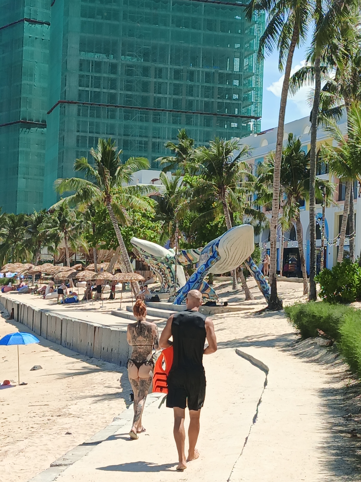 In an outdoor scene, a couple walks away from the camera on a sandy pathway. The man, on the right, wears a black tank top and black shorts, holding an orange life vest. The woman, on the left, has a heavily tattooed back and wears a black bikini. They are walking on a beach with palm trees and umbrellas in the background. A large, colorful whale sculpture is visible to the left of the pathway. Behind the beach, a multi-story building covered in green netting, likely under construction, dominates the upper portion of the image. The environment appears to be a tropical resort area.