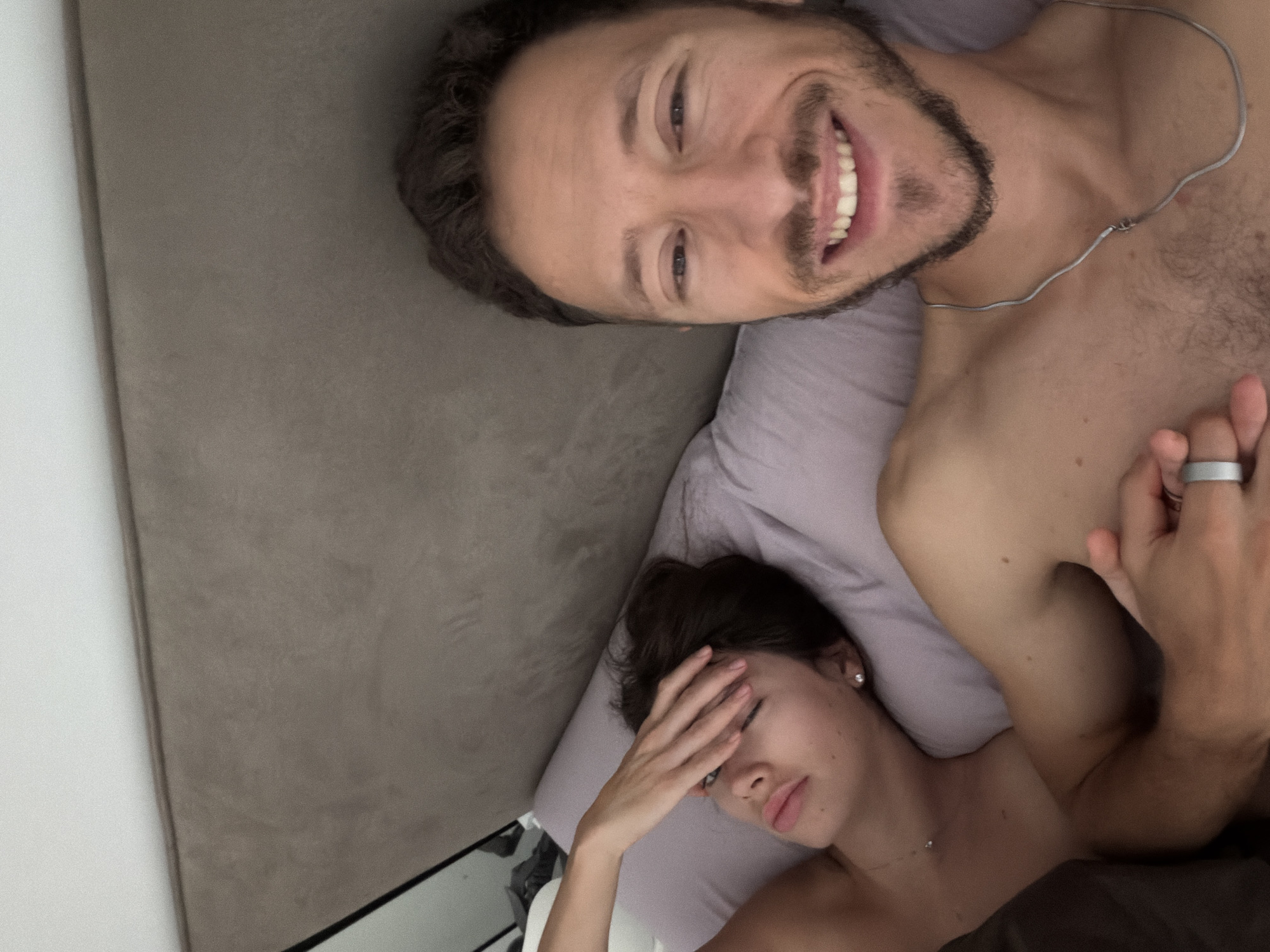 A top-down shot depicts a man and a woman lying in bed. The man is smiling towards the camera and wearing a silver chain necklace. The woman is next to him with her hand covering her eyes. The scene is inside a bedroom. It's impossible to determine the location specifically to Tel-Aviv, Israel.