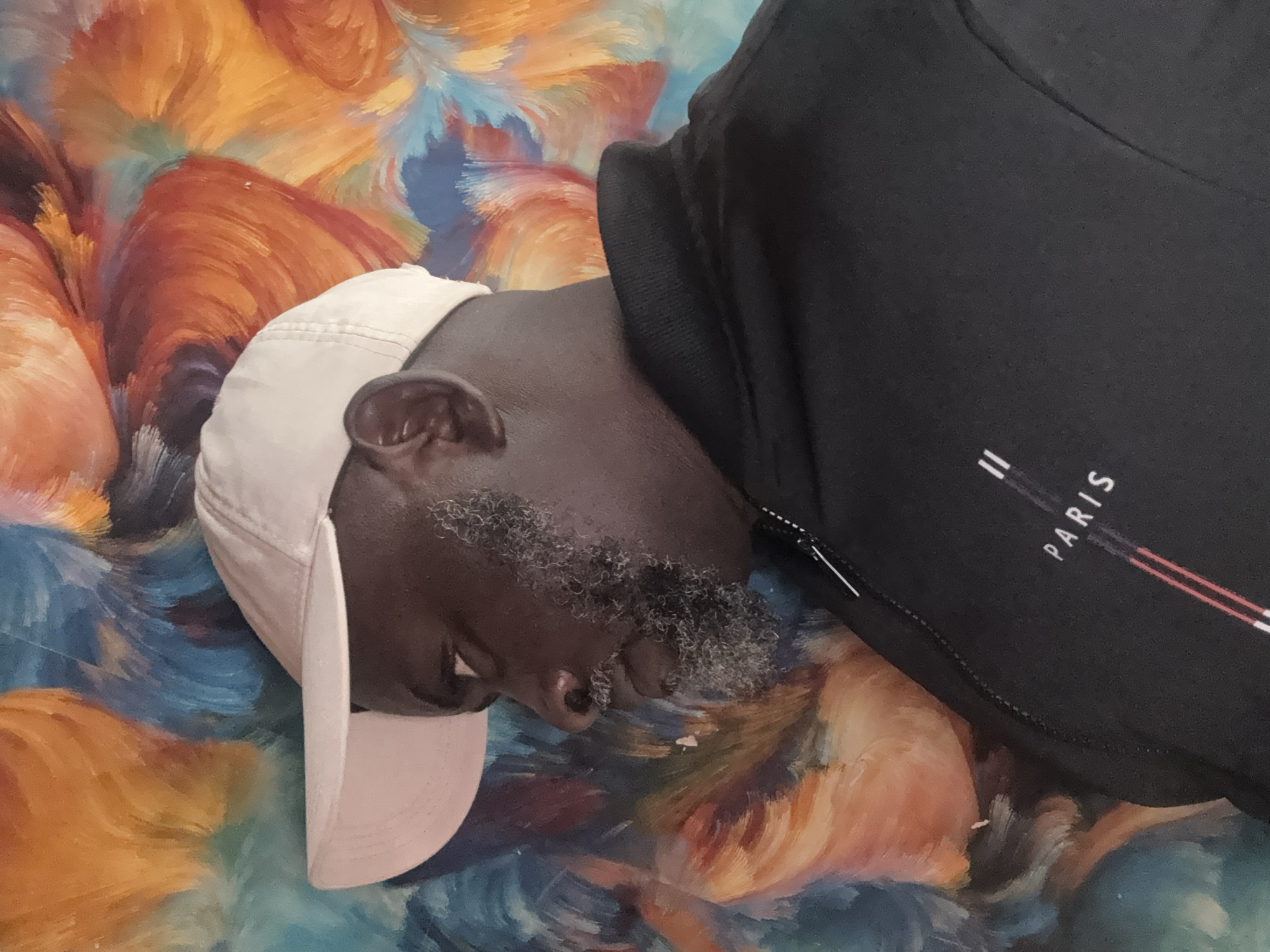 A medium shot captures a Black man, seen in profile facing left, against a backdrop of a vibrant, abstract painting. He wears a light-colored baseball cap and a black jacket with a zipper and the word "PARIS" printed on it in white lettering, accompanied by red and white stripes. His beard is graying. The background painting features swirling brushstrokes in shades of orange, blue, and yellow. The image does not provide any information that can locate it in Ado, Nigeria.