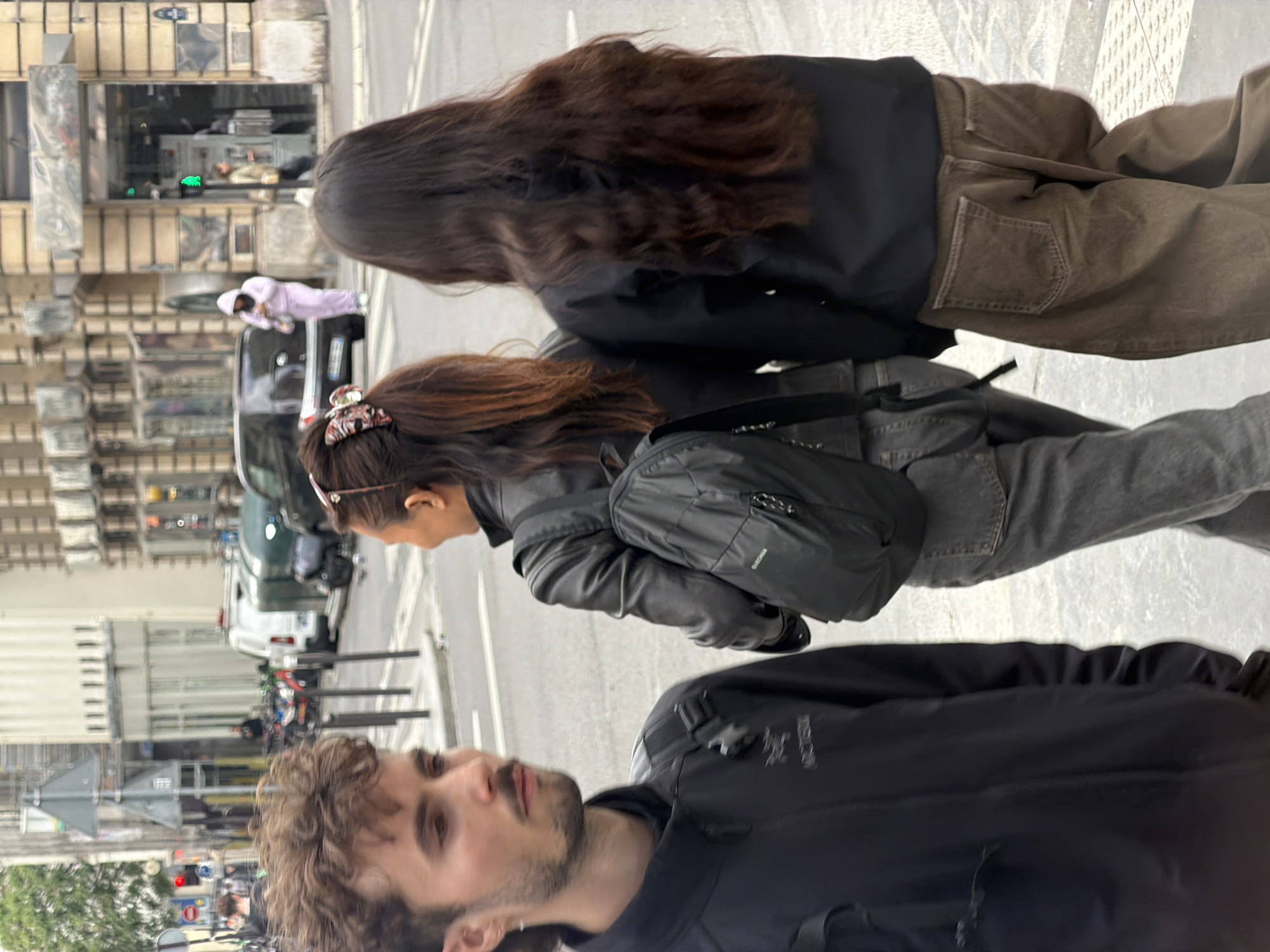 The image captures an urban street scene in Paris, viewed from a slightly elevated perspective. In the foreground, a man with curly brown hair and a beard is partially visible, looking towards the left side of the frame. He is wearing a black jacket, with the brand name "Arc'teryx" discernible on his shoulder.
Behind the man, two women are walking ahead. The woman closer to the foreground has light brown hair tied back with a patterned scrunchie. She wears a black jacket, grey jeans, and a black cross-body bag with "Quechua" written on it. Further back, another woman with long, dark brown wavy hair is seen from behind, wearing a black jacket and brown cargo pants.
The background reveals a bustling city street lined with multi-story buildings of stone or concrete, typical of Parisian architecture. Numerous vehicles are present, including a black car, a white van, and several parked motorcycles or scooters. In the middle distance, a person dressed in a light pink tracksuit is walking on the sidewalk next to a parked car. Traffic lights and various street signs are also visible, contributing to the urban setting. The lighting appears soft and diffused, suggesting an overcast daytime.
