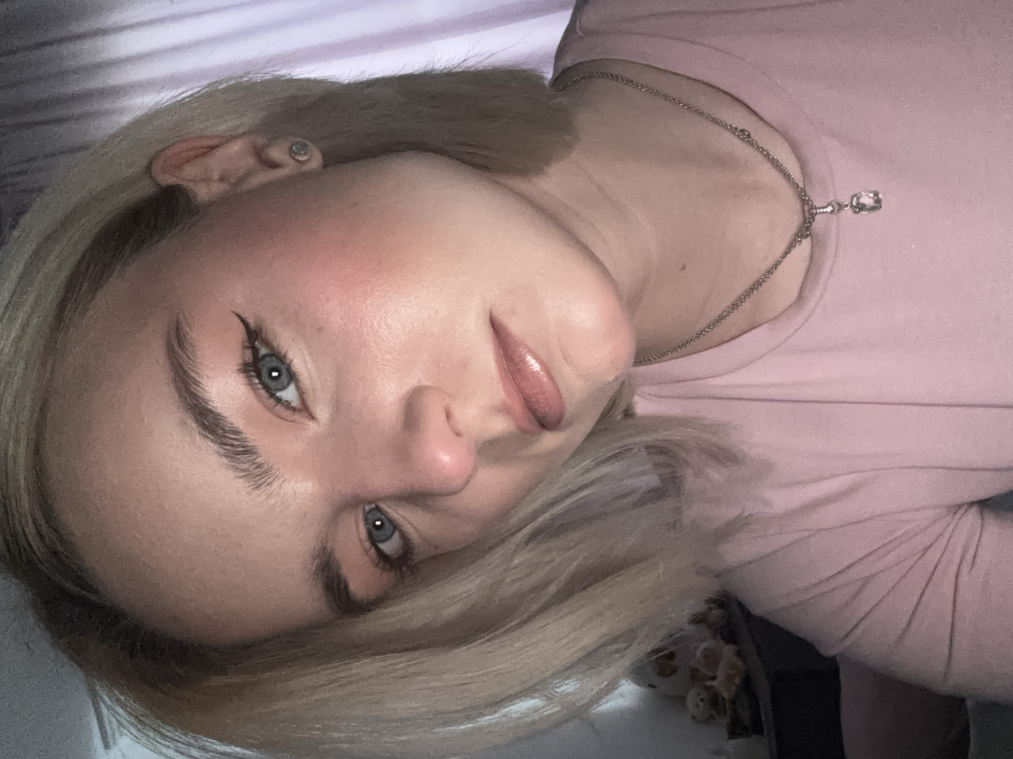 A young, fair-skinned woman with blonde hair and blue eyes is pictured from a close-up perspective. She is wearing a light pink shirt and a silver necklace with a pendant. The woman's face is tilted, and her makeup includes eyeliner and blush. A small earring is visible in her ear. The background appears to be a light-colored surface, possibly fabric. The location is unspecified.