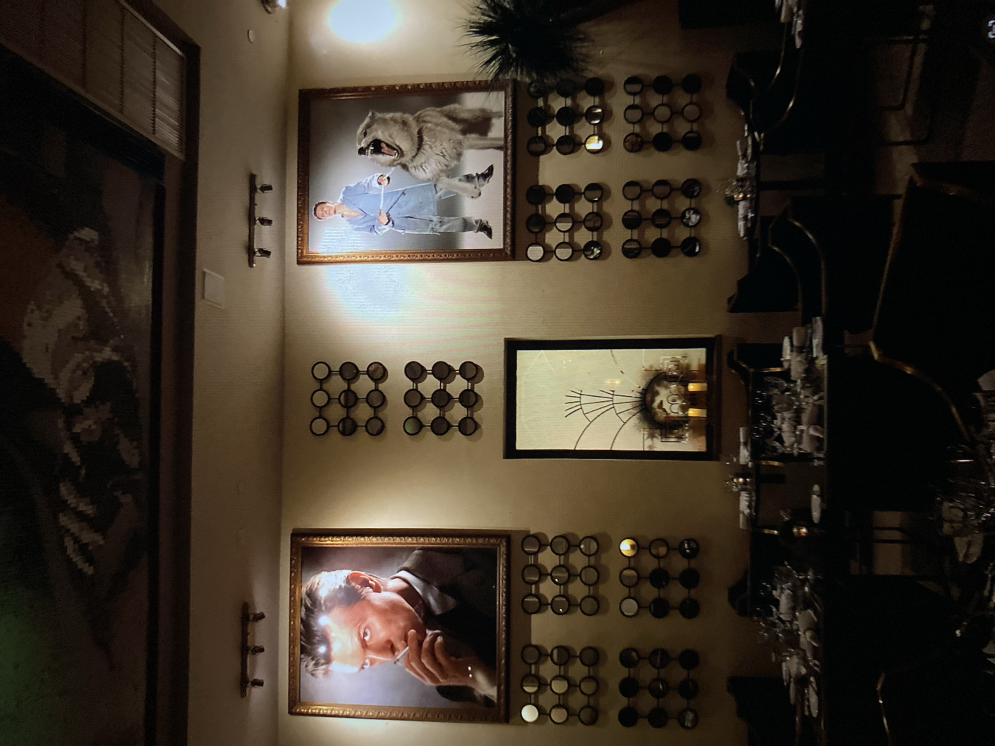 The image depicts an indoor scene, likely a dining or living area, with a wall adorned with various artworks and decorative elements. The primary subjects are framed pictures and decorative wall hangings.
One prominent framed picture features a man in a light blue suit, holding a measuring tape, with a large wolf in the background. The wolf appears to be mid-snarl, with its mouth open. Another framed picture shows a close-up of a man's face, looking intently towards the viewer while holding a cigarette to his lips. A third framed artwork, positioned above the man with the cigarette, appears abstract with circular elements and a central, perhaps clock-like, motif.
The wall is also decorated with several grid-like arrangements of circular mirrored objects, creating a repeating pattern. Two sets of these circular decorations are visible, one above the man with the cigarette and another to the right of the picture of the man and wolf. There are also a few small, dark sconces mounted on the wall.
To the left of the central artwork, a large, dark, framed object is partially visible, showing a textured pattern. In the background, towards the right side of the image, tables with glassware and what appear to be place settings are visible, suggesting a dining or event space. The lighting is dim, with spotlights illuminating the artwork, creating highlights and shadows. The overall mood is somewhat dramatic and artistic due to the bold imagery and the interplay of light and shadow. No text is readily visible in the image.