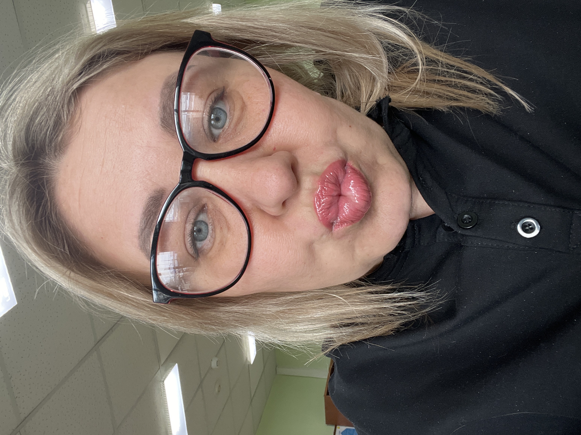 A woman with blonde hair and blue eyes is wearing black-rimmed glasses and a black collared shirt with buttons. She is pursing her lips in a pouting expression. The background shows a light-colored ceiling with fluorescent lights, suggesting an indoor location like an office or institutional building. Although the location is given as Chaikovsky, Russia, there is no direct visual information in the image to confirm this.
