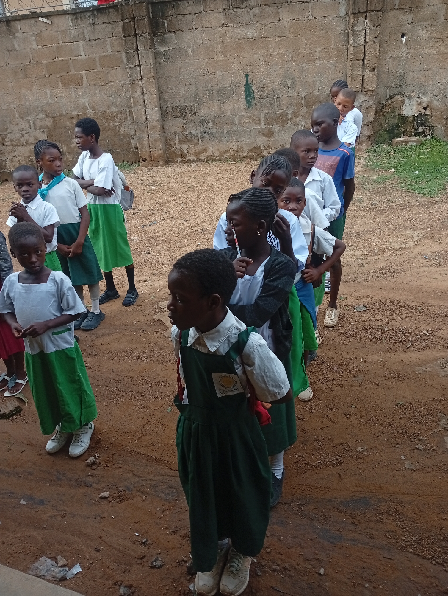 In Narayi, Nigeria, a group of children, likely students based on their matching uniforms of white shirts and green skirts or pants, stand in a somewhat disorganized line on a reddish-brown dirt surface. The uniforms vary, with some children wearing white tops with green skirts and others in dark green overalls over white shirts. Footwear ranges from sneakers to sandals. The children appear to be of African descent.
Behind the children is a weathered, concrete-block wall with some discoloration. Above the wall is a fence of barbed wire. The ground has a mixture of dirt, small rocks, and some debris. A few children are looking towards the left of the frame, while others are looking forward. The lighting appears to be overcast or indirect.