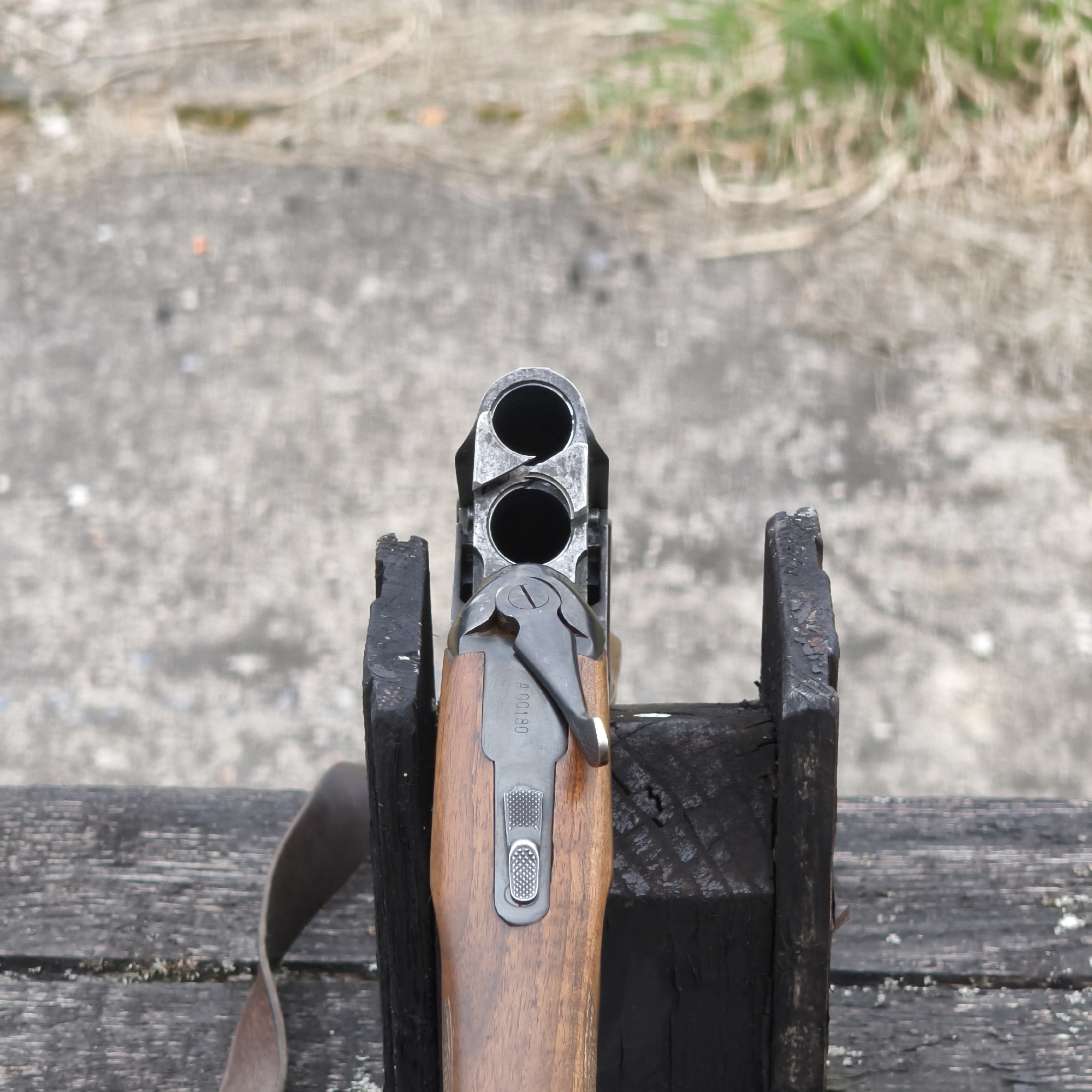 An over-under shotgun is prominently featured in a close-up, eye-level shot, resting vertically in a dark, weathered wooden support. The shotgun's breach is open, revealing the two circular barrel openings. The firearm has a rich, brown wooden stock, and its metallic components, including the receiver and part of the firing mechanism, are visible. Etched onto the metal surface below the opening lever, the serial number "A 00180" can be clearly seen. A textured, possibly rubberized, button and a latch mechanism are also present on the top of the receiver. A dark brown strap, resembling a rifle sling, is visible on the wooden surface to the left of the stand.
The background is blurred, indicating an outdoor setting with a shallow depth of field. It consists of a grey, textured ground surface, possibly concrete or gravel, transitioning into an area with sparse, light-colored grass and dried vegetation. The lighting is natural and diffuse, suggesting it is daytime, potentially under an overcast sky. No human figures are present. The scene likely takes place at an outdoor shooting range or a similar practice area, with the shotgun either being prepared for use, unloaded, or temporarily stored. This location context is associated with Парэцкі сельскі Савет, Belarus.