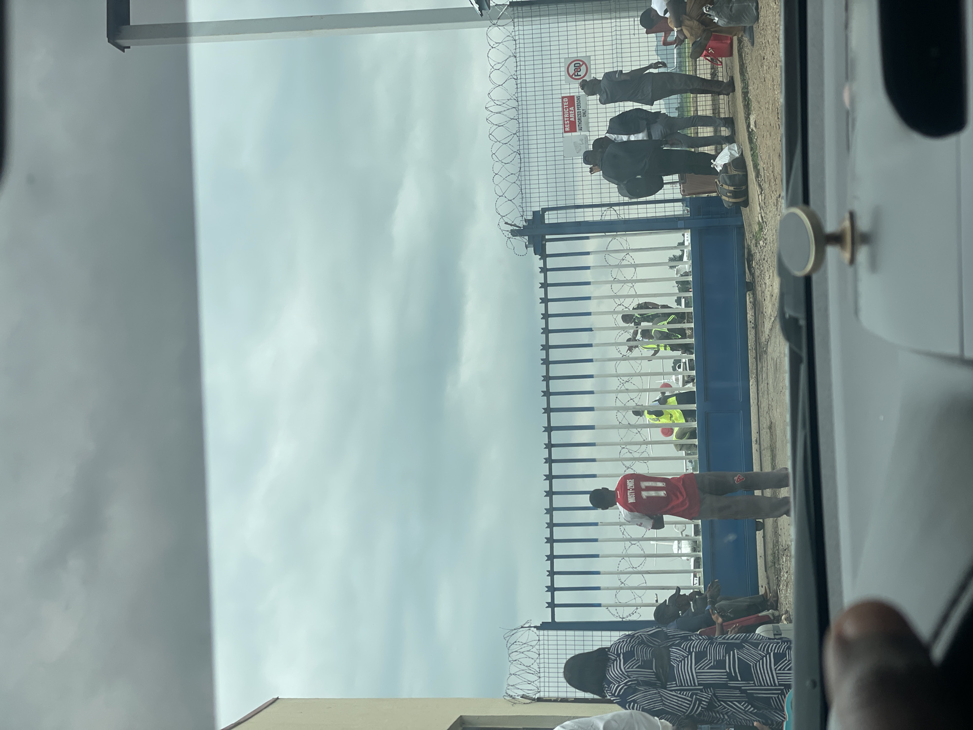 The image depicts a group of people gathered near a blue metal gate topped with barbed wire, likely at an airport or airfield in Gwaska, Nigeria. The sky is overcast with dark clouds. On the left, a person wearing a patterned dress is seen from behind, and next to them, a man in a red jersey with the number 11 and the name "MUSTY-2-NIKE" is standing. Several individuals in high-visibility vests are also visible near the gate, suggesting they are security or airport personnel. Some people are carrying luggage. In the background, small aircraft can be seen. There is a "RESTRICTED AREA AUTHORIZED PERSONS ONLY" sign on the fence, as well as a circular sign with "FOD". The scene appears to be an entrance or checkpoint.