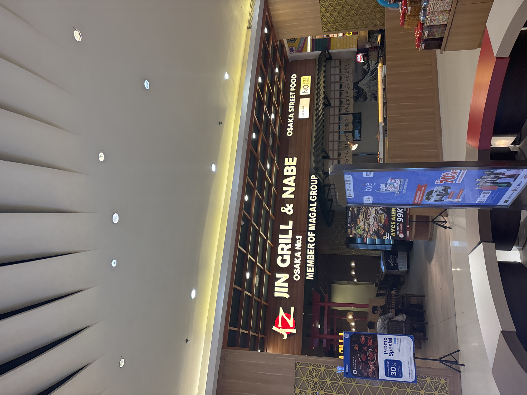The image depicts the exterior of a Japanese restaurant named "JIN GRILL & Nabe Osaka Street Food" located in North Jakarta, Indonesia. The restaurant has a prominent wooden facade with a large illuminated sign. Below the main sign, a smaller sign reads "MEMBER OF MAGAL GROUP." The restaurant appears to be situated within a mall or commercial complex, as evidenced by the lighting fixtures in the ceiling and the polished floor. To the left of the restaurant entrance, there is a promotional poster advertising "Promo Spesial 30% Cashback." To the right, another promotional display features a car and mentions "UP TO 70% OFF." Several customers are visible inside the restaurant, seated at tables and interacting with staff. The overall environment suggests a busy and inviting dining establishment.