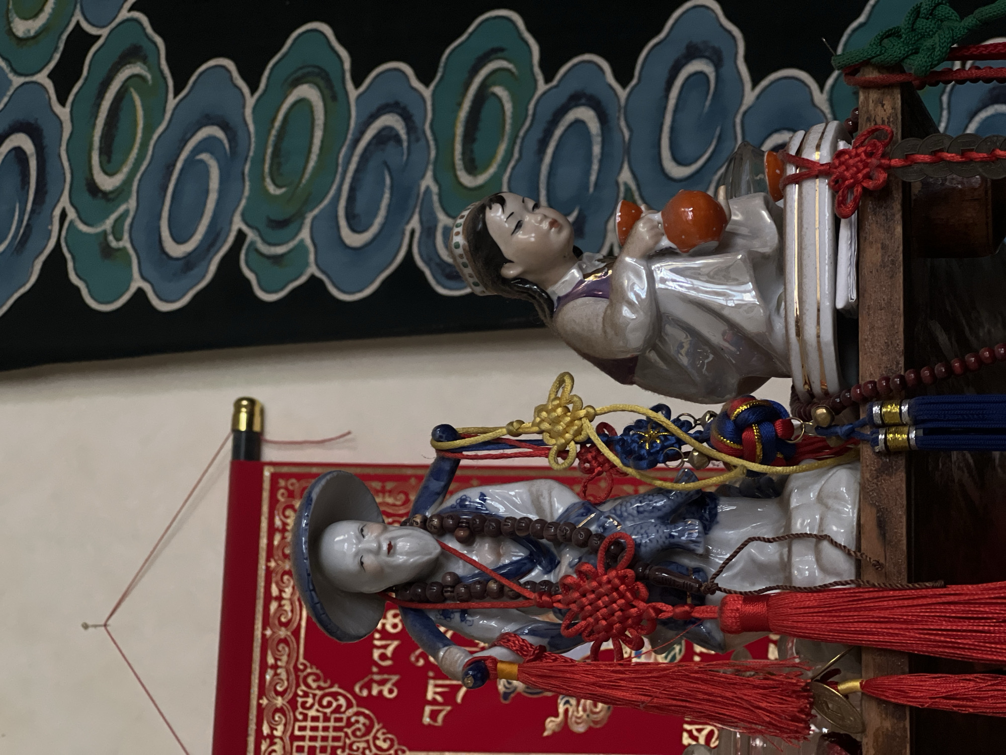 This image displays two intricately detailed ceramic figurines positioned vertically against a decorative background, suggesting an indoor display setting.
The upper figurine depicts a female figure with a serene expression, dark hair, and a patterned headpiece. She wears a light-colored, possibly pearlescent, garment with purple accents and holds two small, spherical orange objects, possibly rattles or instruments. She is adorned with red knotted cords and has small metal coins attached to a wooden rod beside her.
Below her, the second figurine portrays a male figure with a calm expression, wearing a wide-brimmed, grey-blue hat and a blue and white patterned robe. A long strand of dark, spherical beads is draped around his neck. This figure is also elaborately decorated with red, yellow, and blue knotted cords and prominent red tassels hanging below.
The background behind the figurines features a dark fabric or wall covering adorned with a repeating pattern of stylized swirling shapes in shades of blue and teal, resembling clouds or waves. Underneath the lower figurine is a vibrant red textile with intricate gold patterns, possibly a scroll or decorative fabric. There is no discernible activity beyond the static poses of the figurines, and no visible text or explicit location cues within the frame, apart from the decorative elements suggesting East Asian cultural influences.