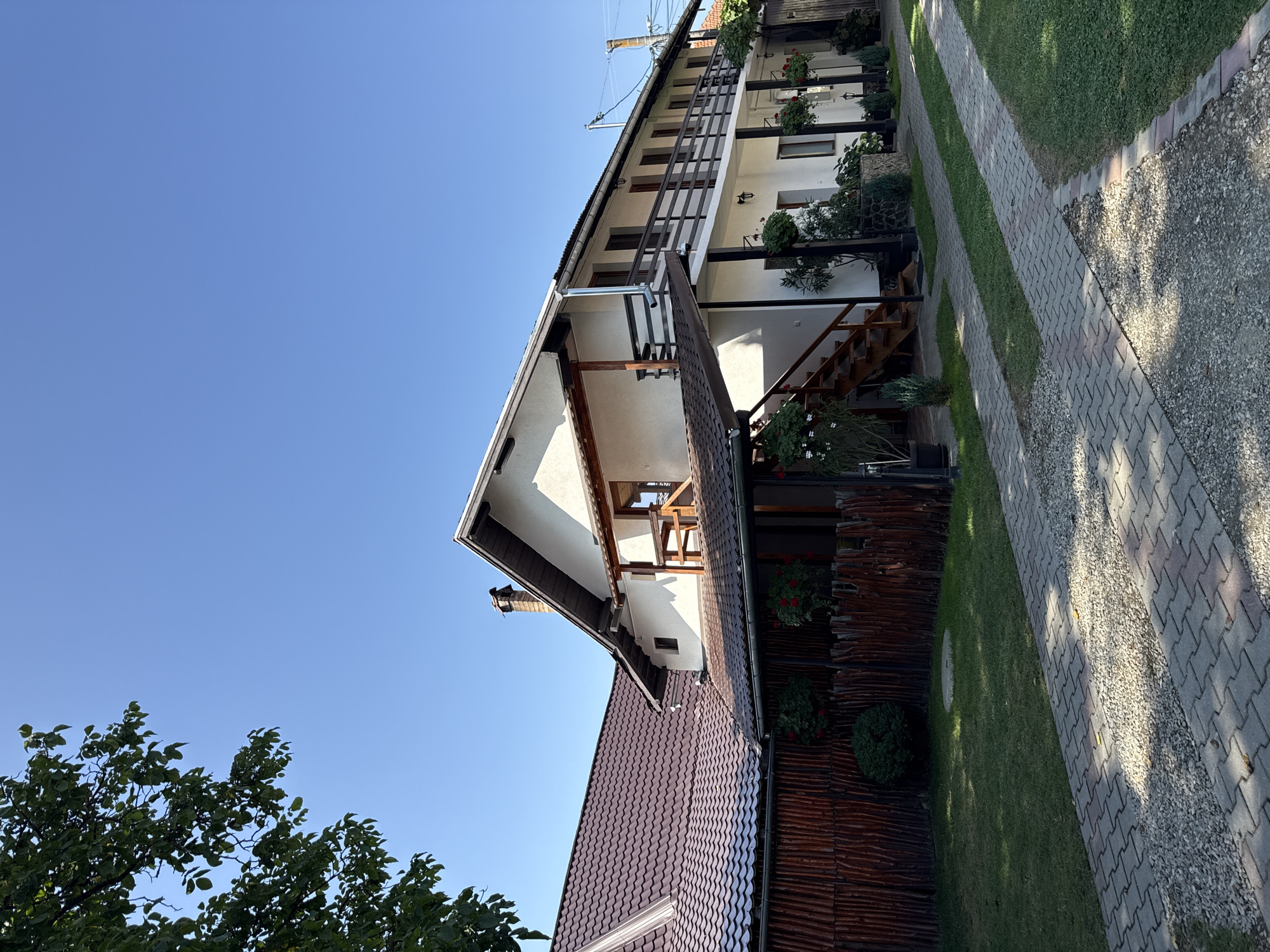 The image depicts the exterior of a two-story building with white walls and dark brown trim, located in Cârțișoara, Romania. The building features multiple windows and a balcony on the upper floor, accessible by an external wooden staircase. Adjacent to it is another building with a brown tiled roof. A gravel and paver driveway runs in front of the structures, bordered by a lawn and a wooden fence. The scene is set against a clear blue sky, with a tree branch visible in the upper left corner.