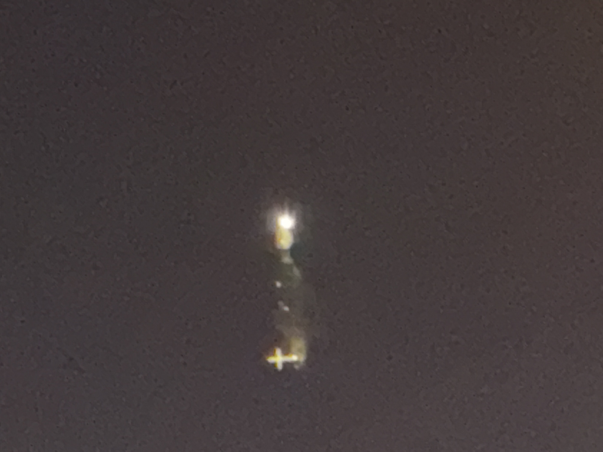 The image is a nighttime photograph taken in Tbilisi, Georgia. It shows a illuminated cross on a hill, with some other lights and what appears to be a structure or trees in the background. The cross is the most prominent feature, glowing yellow and white. The overall scene is dark, with a slightly grainy texture, suggesting the photo was taken in low light conditions. The hill where the cross is located is likely a notable landmark in Tbilisi, possibly a religious site or a memorial.