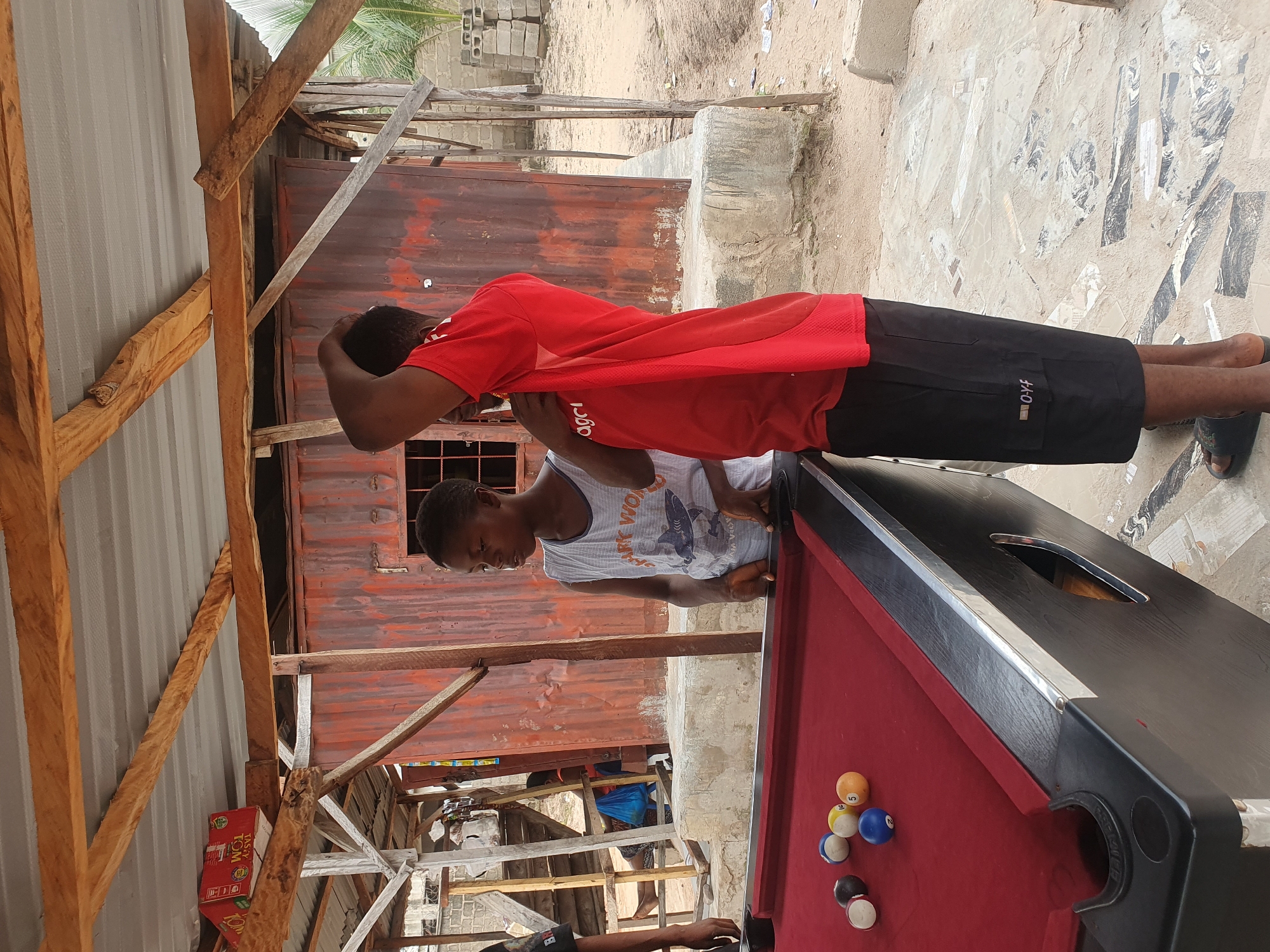 In Lokoja, Nigeria, a scene unfolds around an outdoor pool table under a rustic, corrugated tin roof structure supported by wooden beams. Two young boys are present. One, wearing a white tank top with a graphic design, stands to the right of the pool table, his gaze directed towards it. The other boy, dressed in a red t-shirt and black shorts, stands to his left, his right hand raised to scratch his head. The pool table, covered in red felt, holds a collection of billiard balls, including a blue ball, a yellow ball with the number 5, a white ball with red stripes, a solid black ball, and a white ball with black stripes. Behind the boys, the environment consists of a weathered, reddish-brown corrugated metal building, a concrete base for the pool table, and a sandy ground scattered with debris. The overall setting suggests a casual, outdoor recreational area.