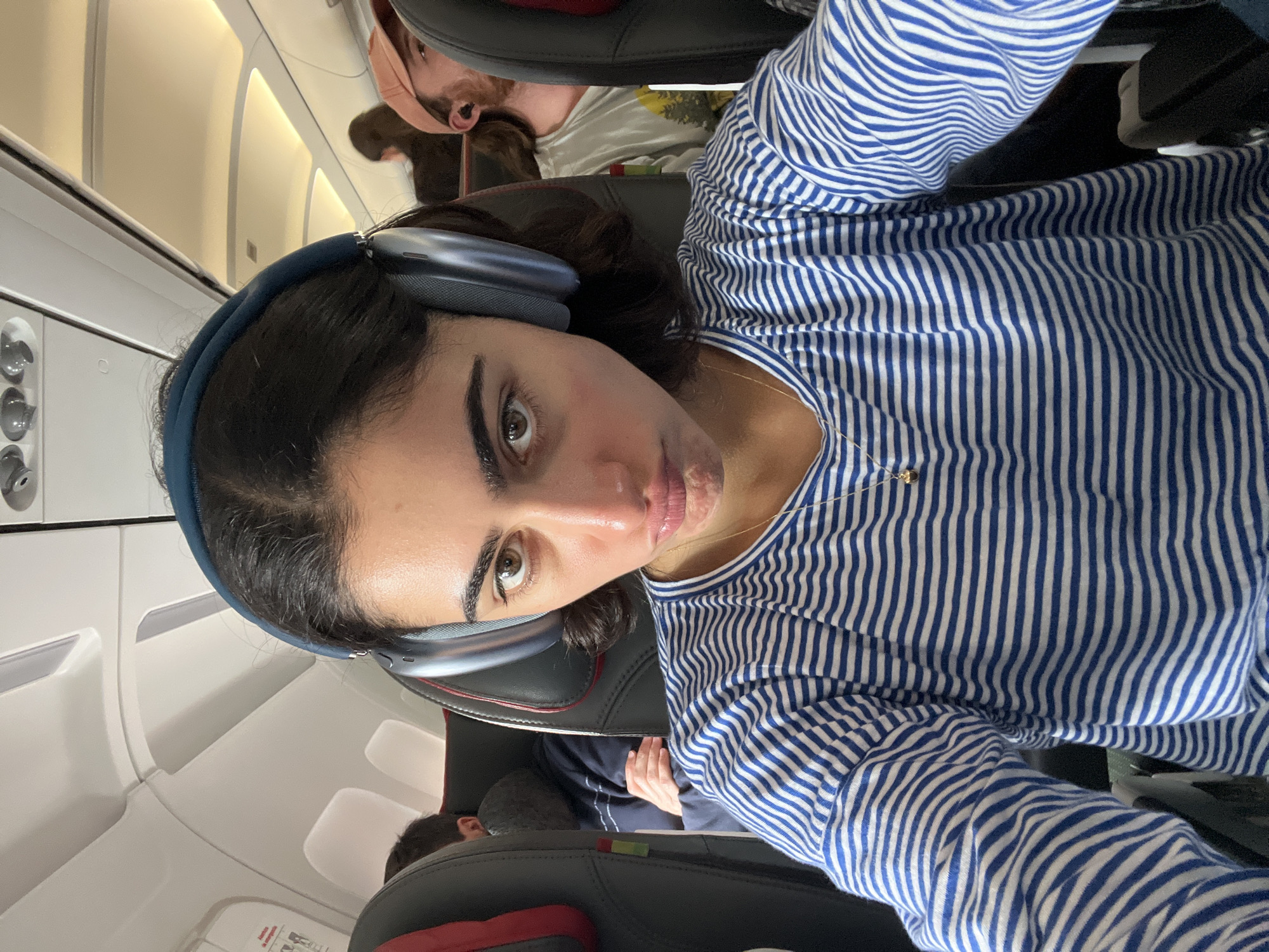 A young woman with dark hair and large headphones is taking a selfie on an airplane. She is wearing a blue and white striped long-sleeved shirt and a gold necklace. Her facial expression is a pout. She is seated in a row of seats, with other passengers visible in the background. The seats are upholstered in black and red. The overhead compartments and lighting of the airplane cabin are also visible. The image appears to have been taken in Paray-Vieille-Poste, France, as indicated by the context of the query.
