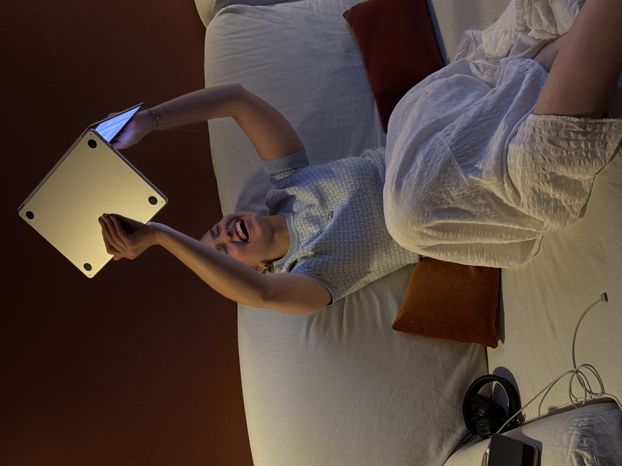 A woman lies on her back on a gray-blue duvet in a dimly lit room. She is holding a silver laptop above her with both hands. The laptop screen is open, revealing a bright glow. She is smiling widely, her mouth open in a laugh. She is wearing a patterned gray-blue short-sleeved shirt and a white dress that is gathered around her. Behind her, there is a red-brown wall. On the duvet, there are two brown pillows. To the right of the duvet, there are black headphones, a phone, and a tangled cable. The location is not clear enough to determine if it is in the city of Milan, Italy.