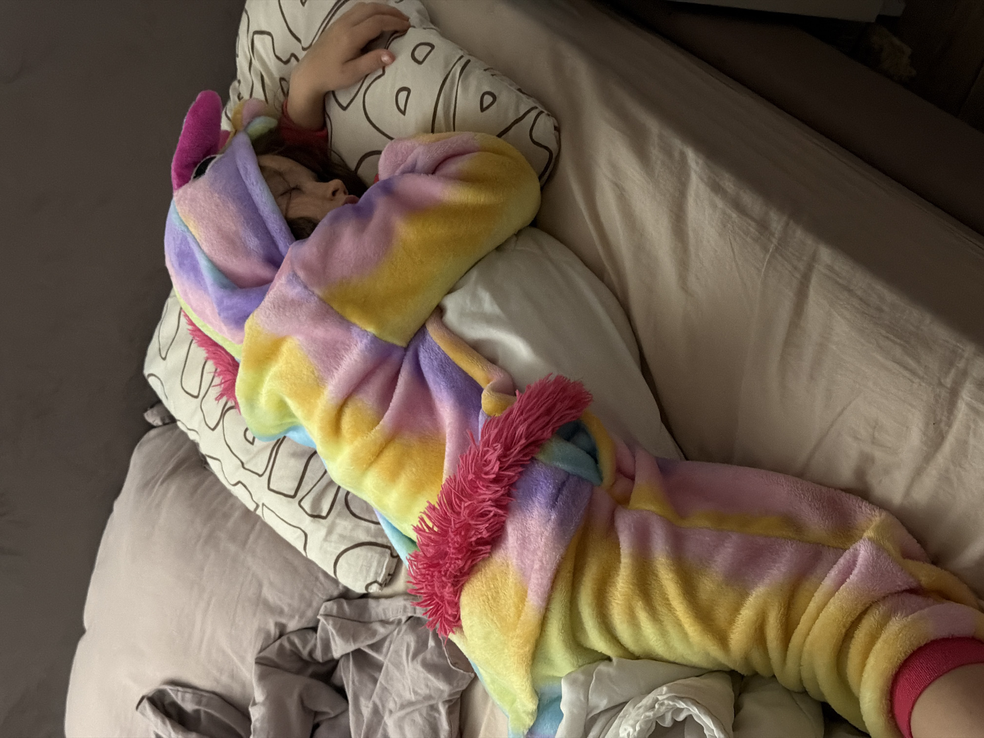 A person wearing a rainbow-colored unicorn onesie is lying in a bed. The person is sleeping, with their face turned to the side. Their right hand is resting on a pillow with a pattern on it. The bed has light-colored sheets and several pillows. The location is unspecified, there is nothing in the picture that suggests it is Perm, Russia.