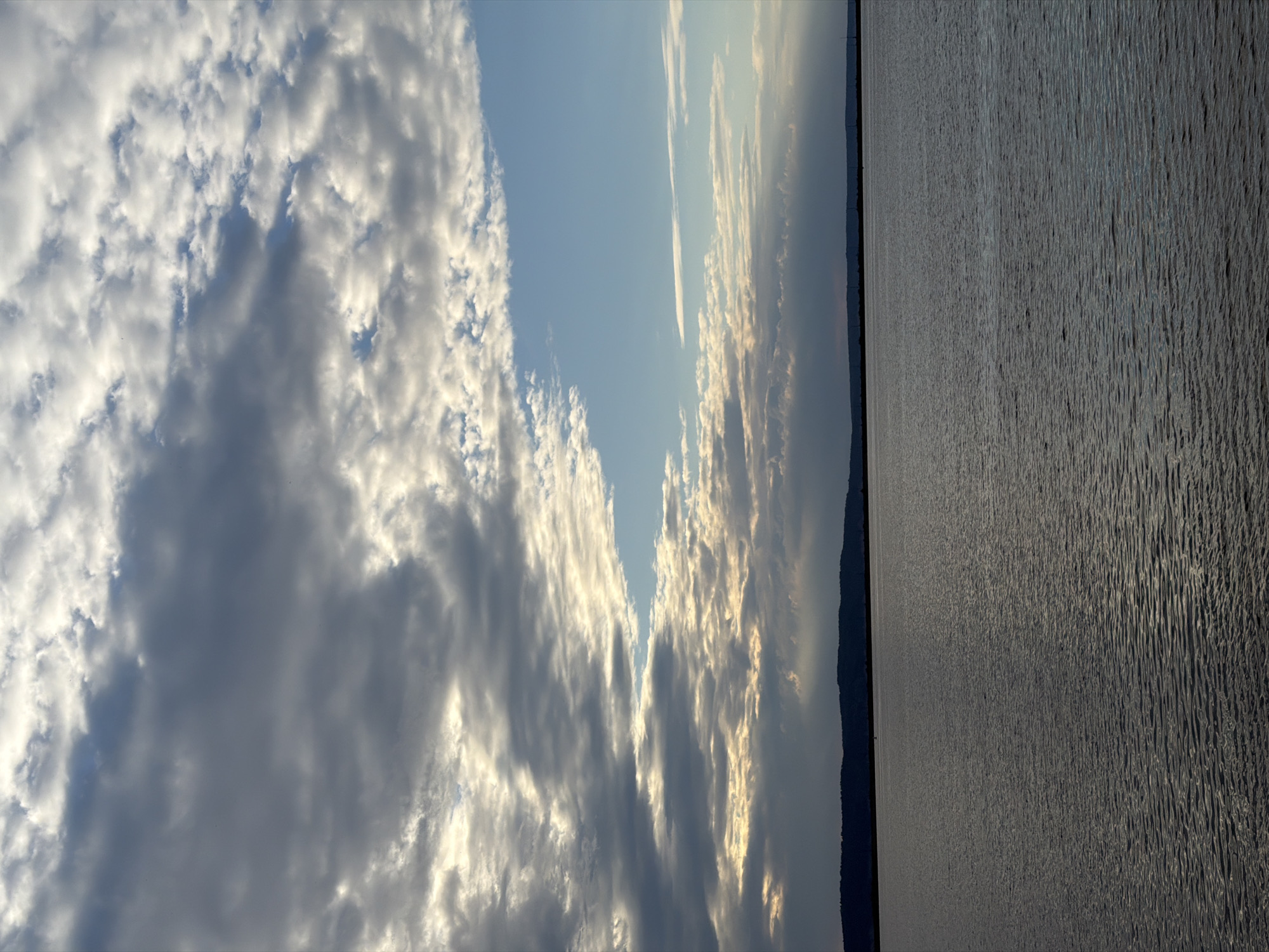 The image depicts a wide expanse of water, likely the Kama River, with a dark, silhouetted shoreline in the distance, characteristic of Chaikovsky, Russia. The sky is dramatic, filled with a mix of dark, heavy clouds and brighter, scattered cumulus clouds, with patches of blue sky visible. Sunlight breaks through in some areas, illuminating the cloud edges and creating a somewhat moody atmosphere. The water is calm with a subtle ripple, reflecting the sky's muted colors. The overall impression is one of a vast, natural landscape under a dynamic sky. There are no people or specific man-made objects visible that would pinpoint the exact location beyond the general geographical context of Chaikovsky.