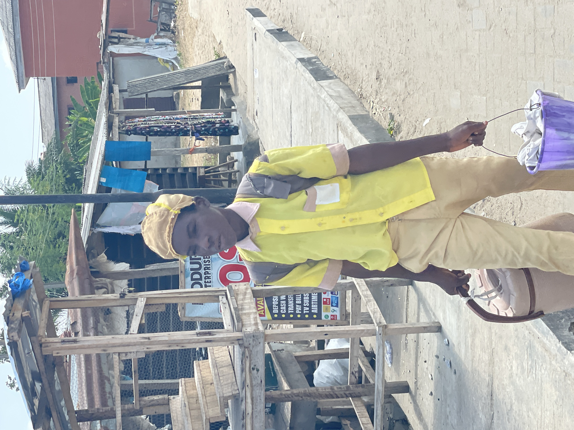 A young man in a yellow uniform and tan pants stands on a concrete curb along a street in Badagry, Nigeria. He carries a purple bucket containing white laundry by its wire handle in his right hand. He wears a tan hat and his head is slightly bowed. Behind him is a makeshift wooden structure with a sign partially visible, reading "ODUF ENTERPRISE". The sign advertises various services including deposits, cash withdrawals, transfers, bill payments, TV subscriptions, and airtime. In the background, other structures, including buildings with corrugated metal roofs and laundry hanging out to dry, are visible. The environment suggests a modest urban or semi-urban area.