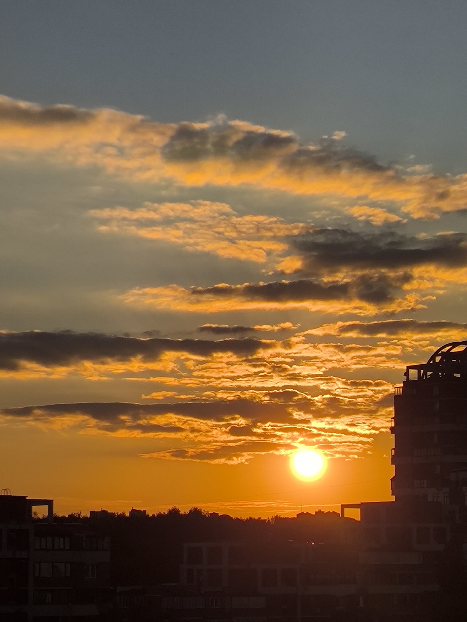 The media file captures a vibrant sunset over the city of Khimki, Russia. The sky is a spectacle of warm hues, with the sun, a bright orb of white and yellow light, low on the horizon, casting a golden glow across the clouds. These clouds, a mix of orange, yellow, and grey, are dramatically lit, with some appearing soft and wispy while others are more defined and textured.
Silhouetted against the intense sky are the outlines of buildings. On the right, a modern high-rise building with distinctive architectural features, including curved elements and balconies, dominates the skyline. To the left and in the foreground, other buildings are visible, appearing as darker shapes against the illuminated sky and landscape. A line of trees forms a dark band between the city buildings and the horizon, suggesting a park or wooded area within or on the outskirts of the city.
The overall scene evokes a sense of tranquility and natural beauty. The time of day is clearly sunset, indicated by the low position of the sun and the dramatic coloration of the sky. The weather appears clear, allowing for an unobstructed view of the sunset. While there are no people visible, the presence of the city buildings anchors the scene in an urban environment. There is no visible text in the media file.