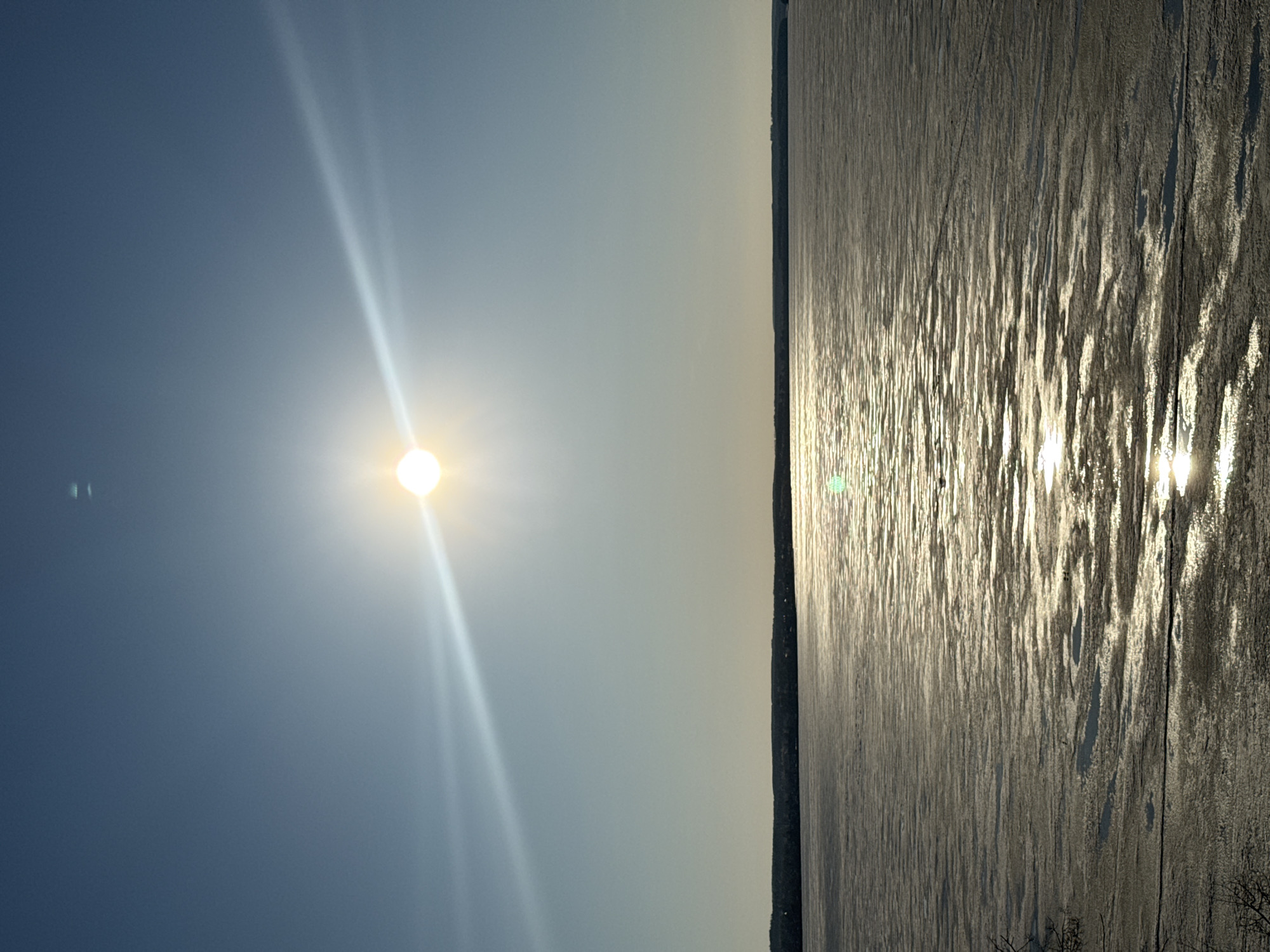 The image depicts a sunny day in Votkinsk, Russia, overlooking the partially frozen Votkinsk Reservoir. The sun is prominent in the sky, with light rays extending downwards. The reservoir surface displays a mix of ice and open water, reflecting the sunlight. The shoreline appears as a thin strip of land separating the sky and the water, with some distant buildings or trees faintly visible. There are no people present in the image.