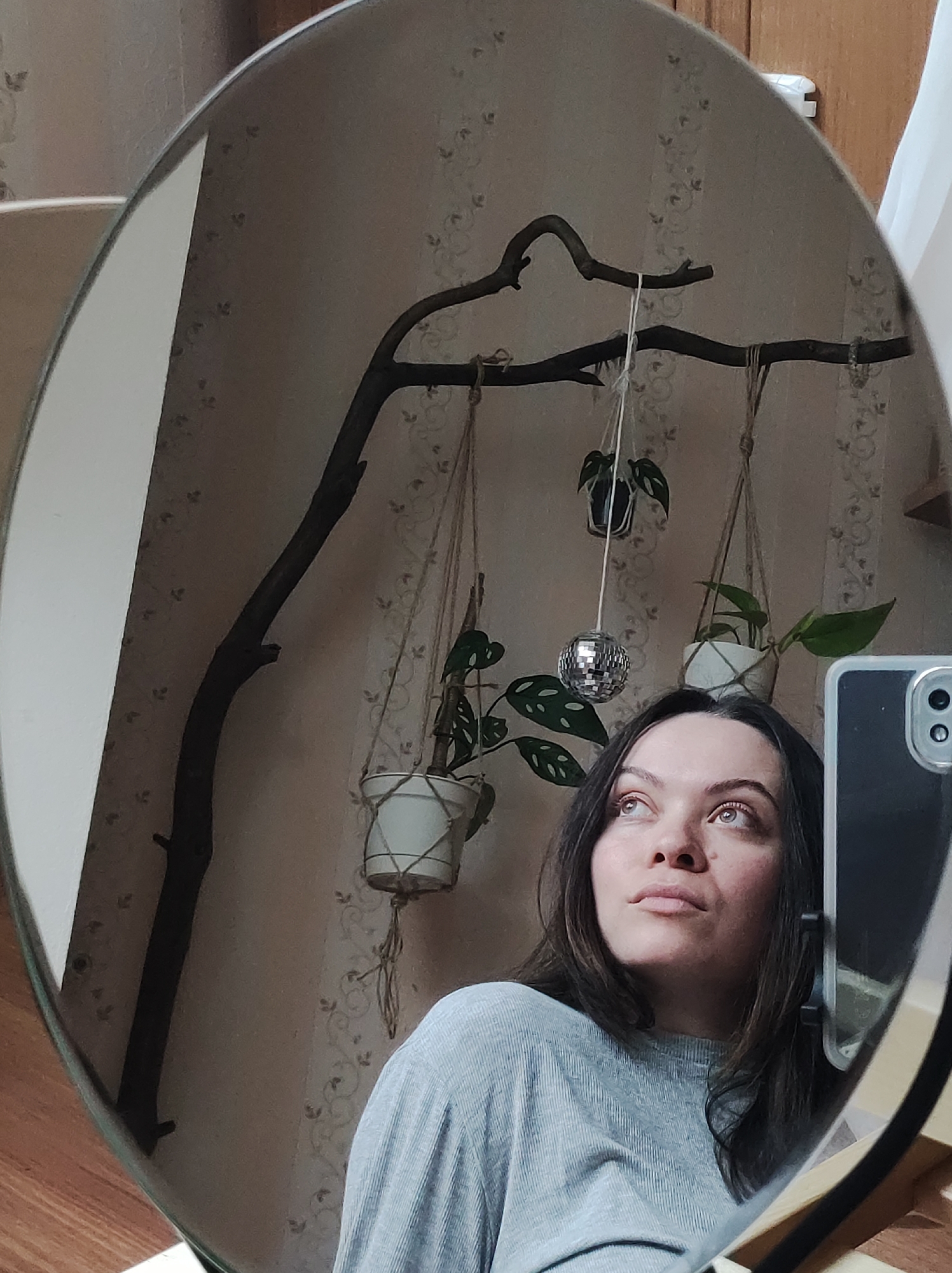 A reflective oval mirror reveals a woman's self-portrait. She is looking upwards with her eyes open. Her shoulder-length dark hair frames her face, and she is wearing a gray long-sleeved shirt. The reflection in the mirror shows a wall covered in patterned wallpaper, above which is an unidentifiable object made out of wood, from which are suspended several small hanging plants in white pots, dangling from ropes. A disco ball hangs near the center of the image. In the foreground, a portion of a cell phone is visible, which is likely being used to capture the image. The scene is indoors. There is no definitive information to confirm the location is Chaikovsky, Russia.
