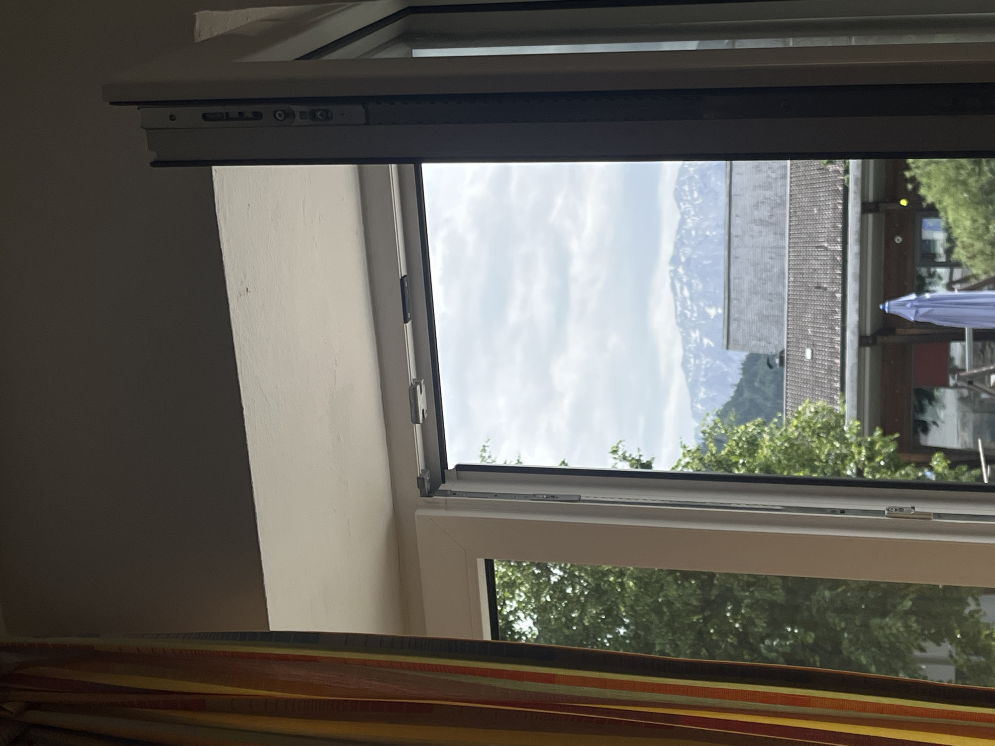 The image shows a view from an open window in Garmisch-Partenkirchen, Germany. The window frame is white, and the window is tilted open at the top. Outside, there is a partial view of a building with a gray tiled roof and snow-capped mountains in the background, partially obscured by clouds. There is a blue umbrella visible. The trees are green. There is a curtain at the bottom of the image.