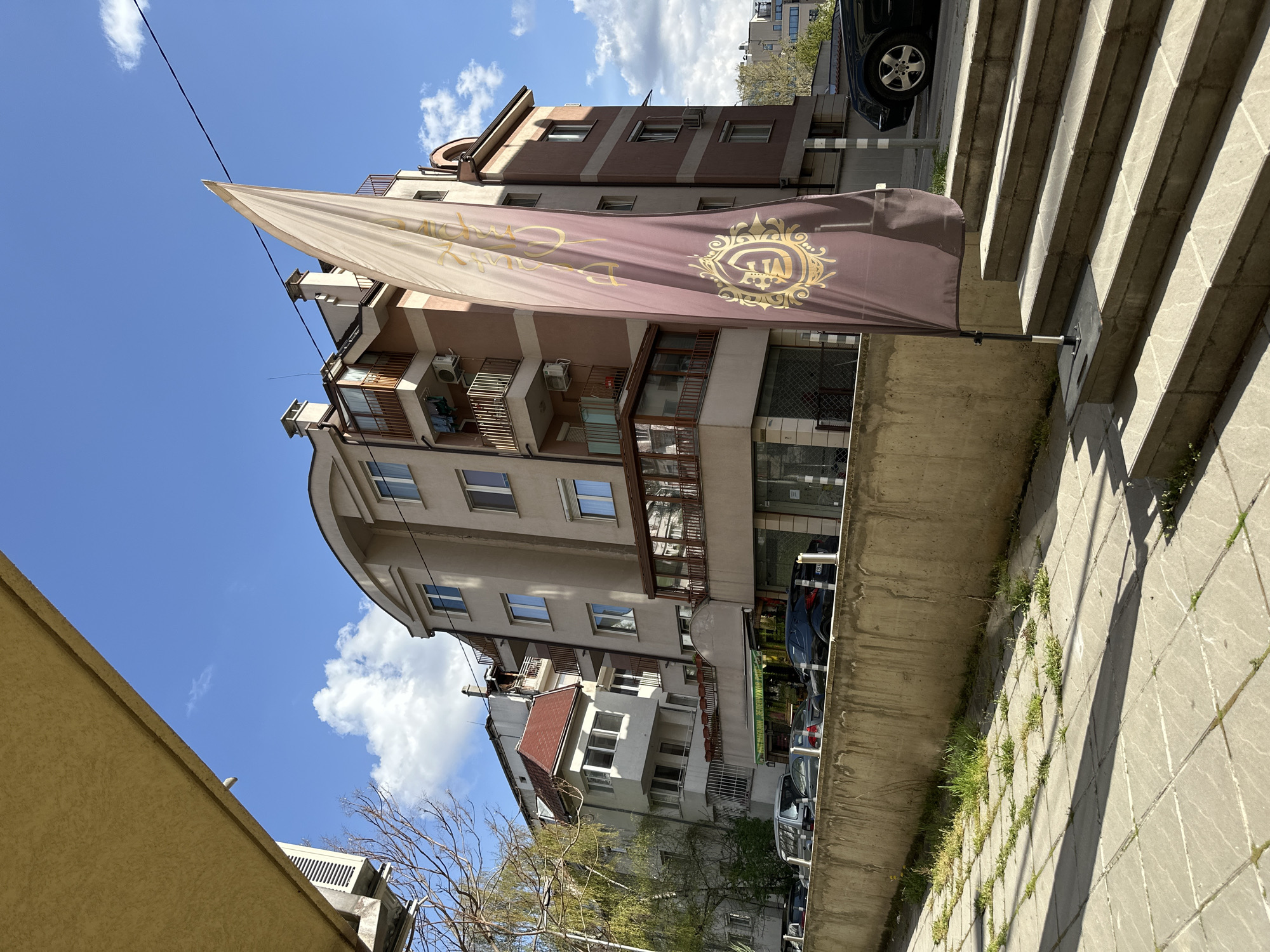 The image, originally rotated 90 degrees clockwise, captures an urban street scene in Sofia, Bulgaria, on a bright, sunny day.
A prominent feature is a large, dark brown flag hanging diagonally from a pole in the mid-ground. The flag is adorned with ornate golden text and a crest-like logo. The logo appears to be a crowned shield emblem, possibly containing the letters "TA." The visible text on the flag, rendered in a decorative script and partially obscured by folds, seems to read "Golden Crown."
Behind the flag, a multi-story building stands, characterized by a light-colored facade with sections of a contrasting reddish-brown. The building features several balconies with metal railings and numerous external air conditioning units. Its architecture suggests a typical European urban structure, possibly residential or mixed-use.
To the right of the flag, a set of concrete stairs ascends, exhibiting strong, distinct shadows indicative of direct sunlight. The paved ground adjacent to the stairs shows patches of green grass growing through the cracks. On the left side, other buildings with red-tiled roofs are visible in the background, alongside bare tree branches, suggesting a season like late winter or early spring. Several parked cars, including a dark-colored vehicle, can be seen along the street. The upper portion of the frame reveals a clear, vibrant blue sky dotted with scattered white cumulus clouds. No people or specific events are depicted, presenting a static street view.