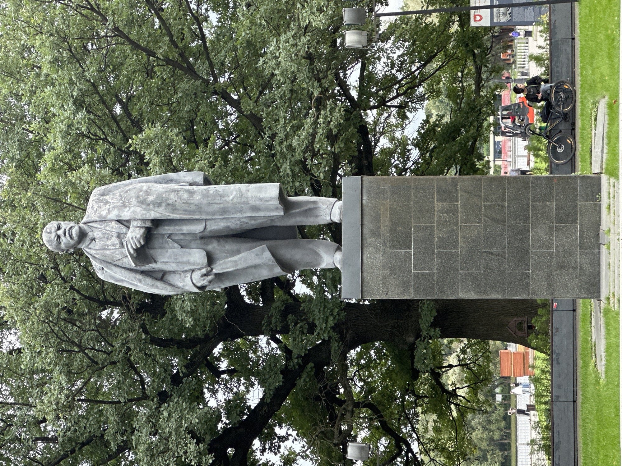 A full-body, gray stone statue of Vladimir Lenin stands prominently on a dark gray stone pedestal in an outdoor park setting. Lenin is depicted wearing a suit and overcoat, holding a book in his left hand. His expression is stern, with his mouth and mustache clearly defined. Behind the statue, a large, mature oak tree with dense green foliage dominates the background. The park appears to be in a public area, with a paved pathway visible behind the pedestal, bordered by a low, dark gray planter. In the distance, several people are walking, and a construction vehicle or forklift can be seen. To the right of the statue, a bicycle is parked next to a person sitting on a low wall, wearing headphones. The ground in the foreground is covered in green grass. The overall impression is one of a monument in a well-maintained, possibly historical, urban park. The statue is located on Lenin Square in Moscow, Russia.