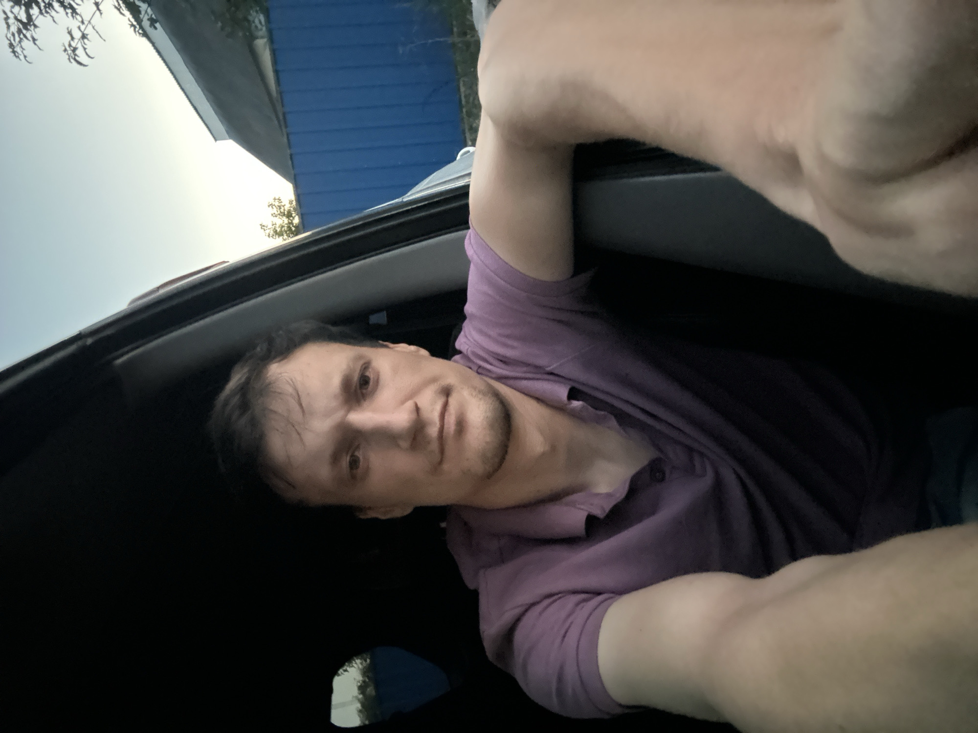 A man is shown from the chest up, sitting inside a vehicle, with his left arm resting on the car door. He is wearing a purple polo shirt and has short, dark hair. His expression is neutral, and he is looking directly at the camera. The background of the image suggests an outdoor setting, with a glimpse of a blue fence, a white building with a sloped roof, and trees visible through the car window. The sky appears clear and possibly shows the early evening light. The location is identified as Shatava, Ukraine.