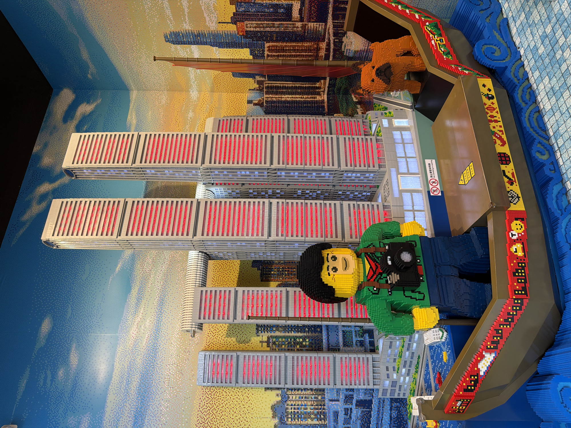 The image depicts a detailed LEGO model of a cityscape, with a prominent boat in the foreground. The cityscape features tall, modern skyscrapers with red and blue striped facades, set against a backdrop of a painted sky with clouds. The boat, also constructed from LEGOs, is filled with blue wave-like pieces simulating water. In the boat, a LEGO figure of a person wearing a green jacket and holding a camera stands to the left. To the right, a brown LEGO dog sits facing the viewer. The boat's exterior is decorated with colorful LEGO patterns and symbols. A sign on the building behind the boat reads "禁止攀爬", meaning "No Climbing". The overall scene is a static, artistic representation of an urban environment, likely a tourist attraction, as the detail and scale suggest it. While the prompt mentions Yuzhong District, China, this specific location cannot be confirmed from the visual information alone; the scene could represent any modern city with similar architecture.