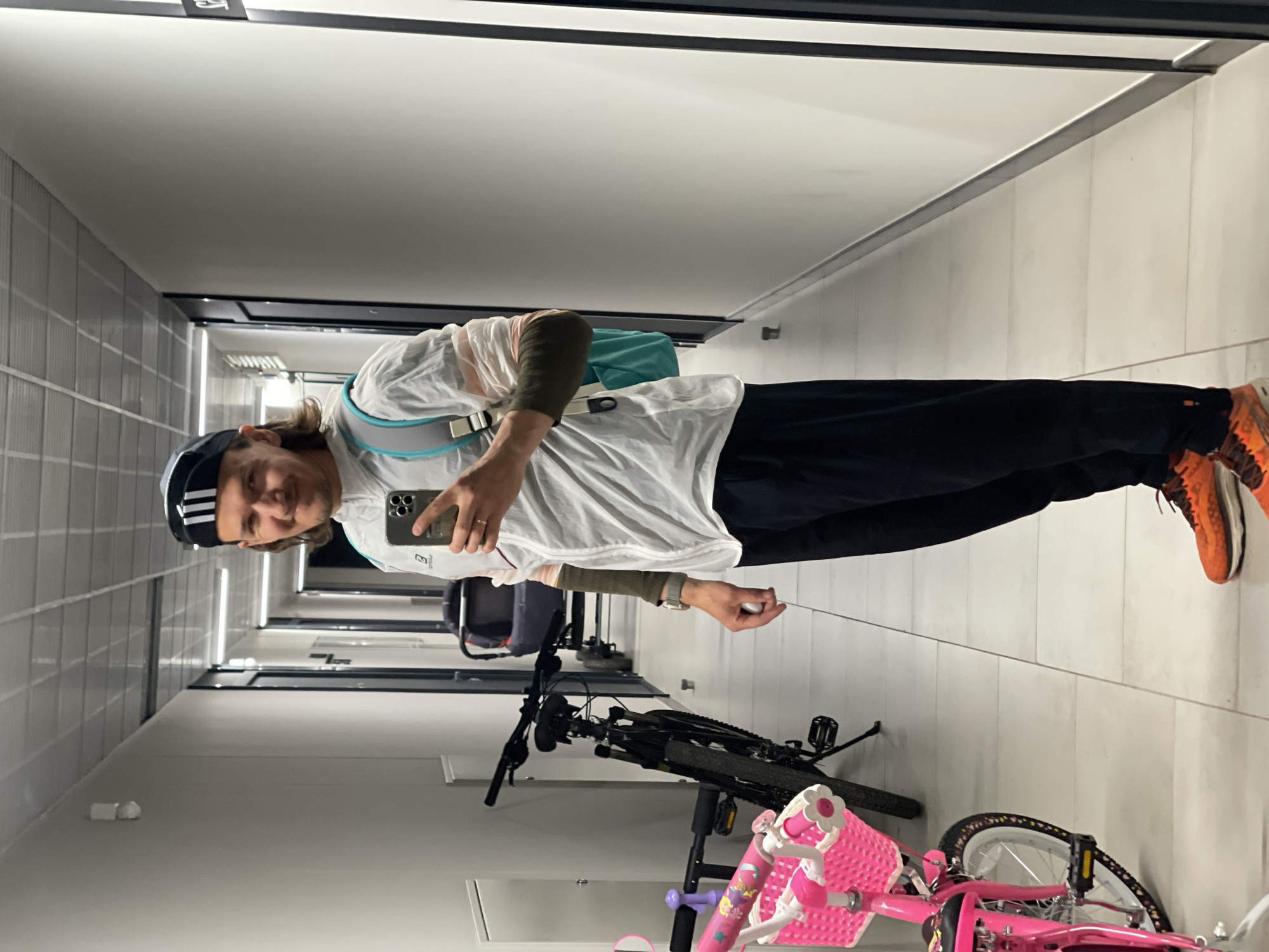 A man stands in a corridor, holding a phone in his right hand, taking a selfie. He wears a black Adidas hat, a white and green long-sleeved shirt, black pants, and orange shoes. A teal bag hangs over his shoulder. Two bicycles are visible behind him: a black mountain bike and a pink child's bike. The corridor has white walls and a gray tiled wall on one side. There are doors and fixtures along the corridor, suggesting an indoor space, possibly an apartment building or similar residential setting. The location is city of Unknown, Unknown.