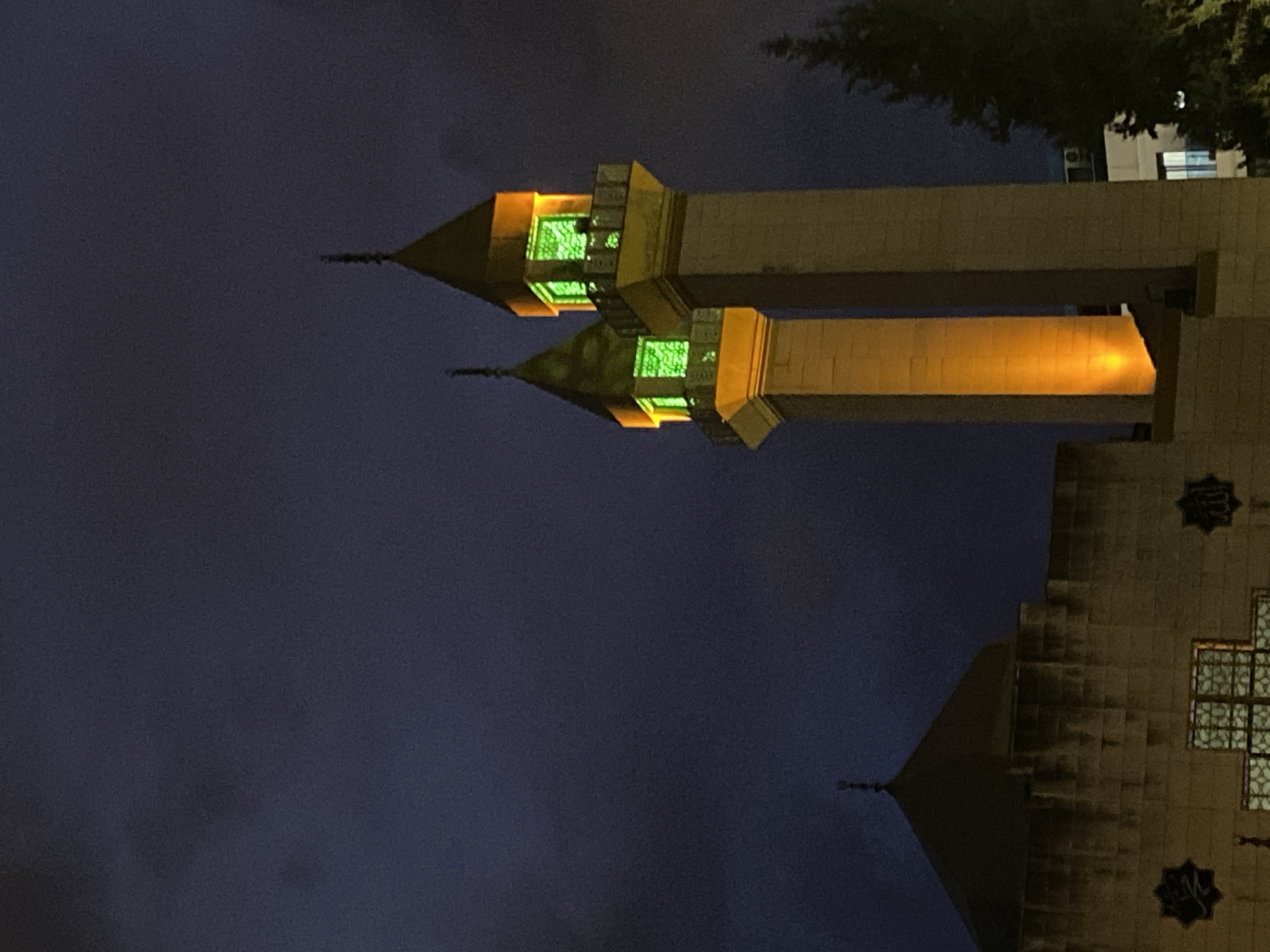 At night, the Alanya State Hospital Mosque in Alanya, Turkey, is illuminated. The mosque's two minarets are visible, each topped with a conical roof and lit by green lights inside. The minarets are connected by a structure with orange lighting. Trees and a building are visible in the background. The sky is dark.