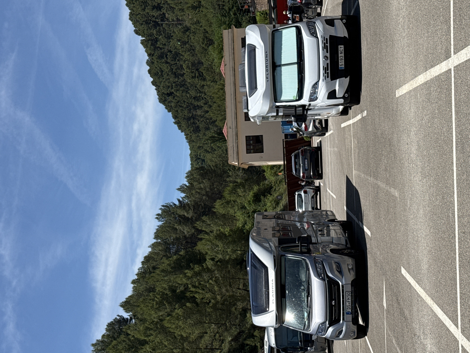 Two recreational vehicles (RVs) are parked in a paved lot. The RV on the left is silver and has a license plate that reads "FDS FH50". The RV on the right is white and has a license plate that reads "5193 NCY". Other vehicles, including cars and possibly another RV, are also visible in the lot, along with a building and a dense forest on a hillside in the background. The sky is blue with scattered white clouds. The location is the parking area for the Cardona Salt Mountain Cultural Park in Cardona, Spain.