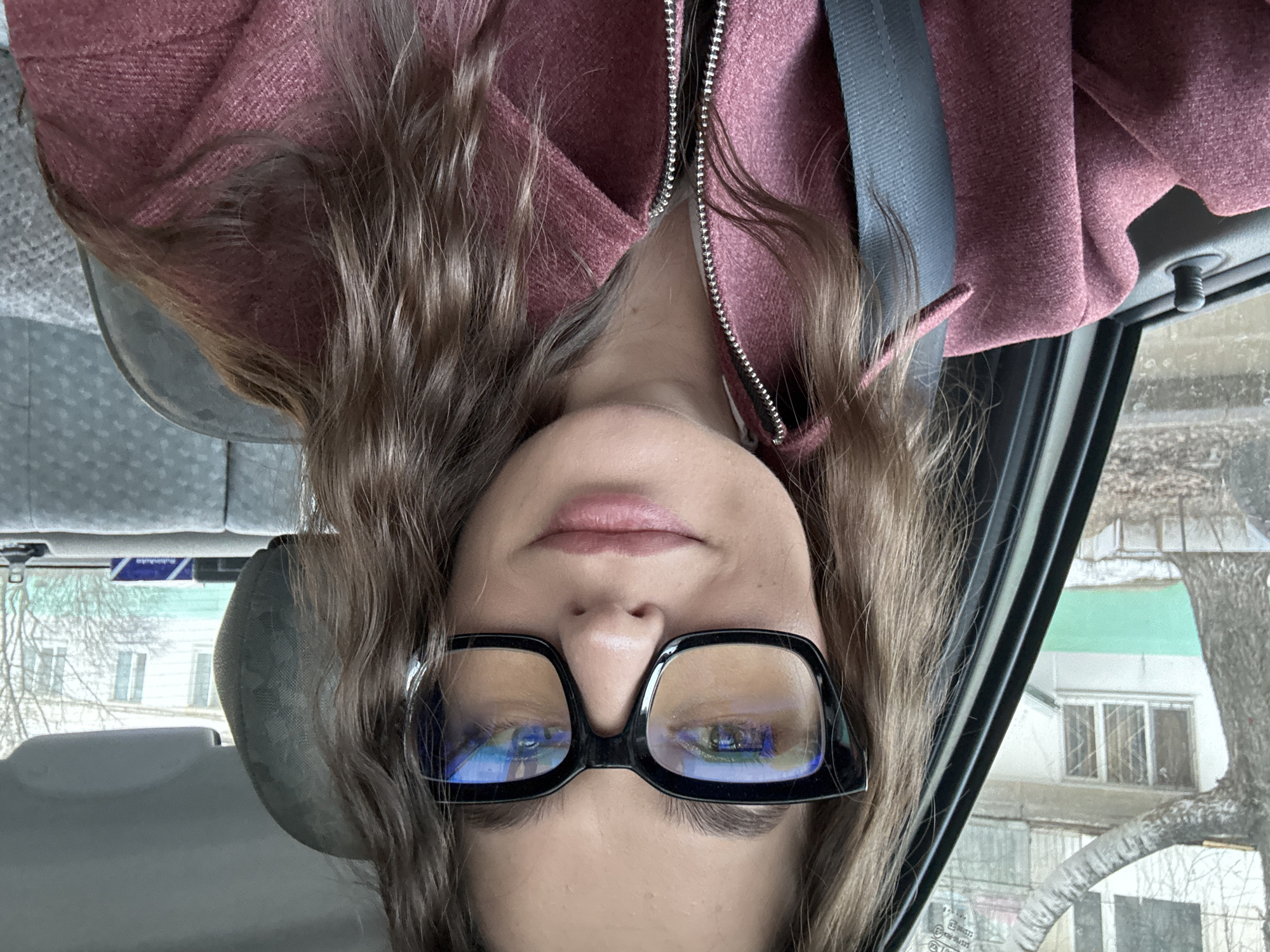 A person with long brown hair wearing glasses and a pink jacket is inside a vehicle. A seatbelt is visible across their chest. The reflection in their glasses suggests a cloudy day. Through the car window, a building with multiple windows can be seen, along with bare trees. The location is consistent with a residential area in the city of Chaikovsky, Russia.