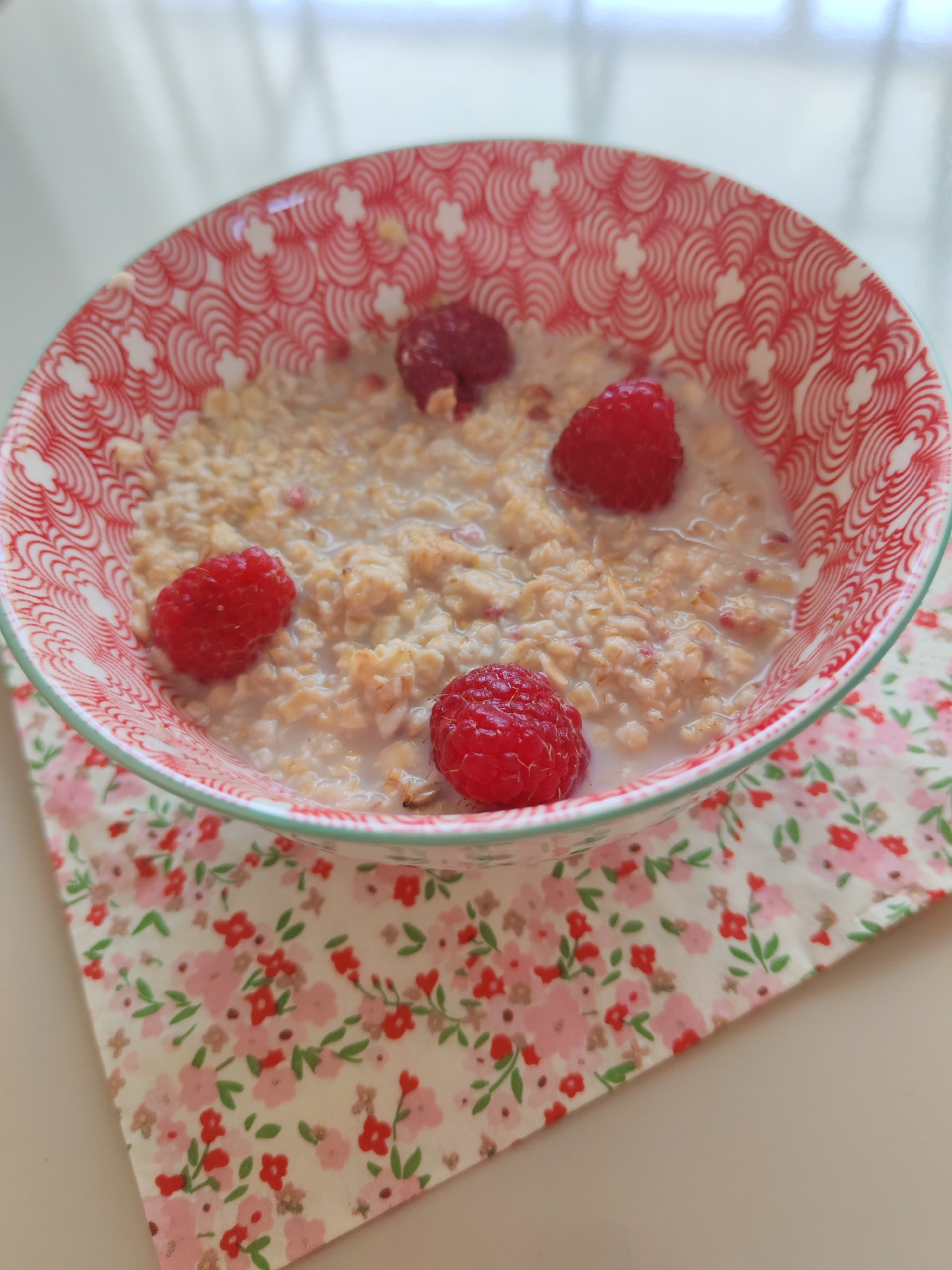 A close-up, overhead shot displays a bowl of oatmeal with raspberries, placed on a floral-patterned cloth. The bowl has a red and white geometric pattern around its rim and contains cooked oatmeal mixed with milk, topped with four whole raspberries. The floral cloth features small pink and red flowers with green leaves on a cream background. The scene is lit by natural light, casting soft shadows. The background is out of focus, showing a light-colored surface. There are no people or animals visible in the image. The scene is static and indoors, likely a breakfast setting. No specific location in Sabunchu Raion, Azerbaijan can be determined from the image.