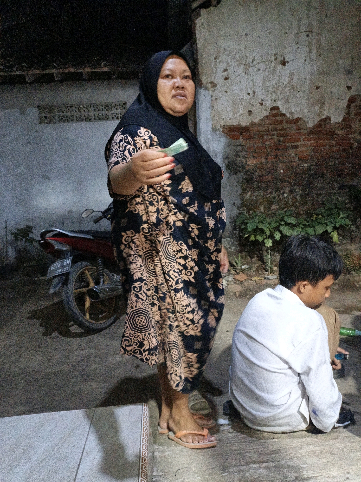 A medium shot captures a street scene in Tanahbaya, Indonesia, at dusk. A plump woman wearing a black hijab and a patterned dress stands in the foreground, holding a small amount of cash. To her right, a young boy in a white shirt sits on the ground, facing away from the camera. A motorcycle is parked behind the woman. The background features a weathered brick wall with exposed mortar, suggesting an unfinished or dilapidated structure. The ground is paved with concrete and dirt. A shadow of a person can be seen on the tiled surface to the left.
