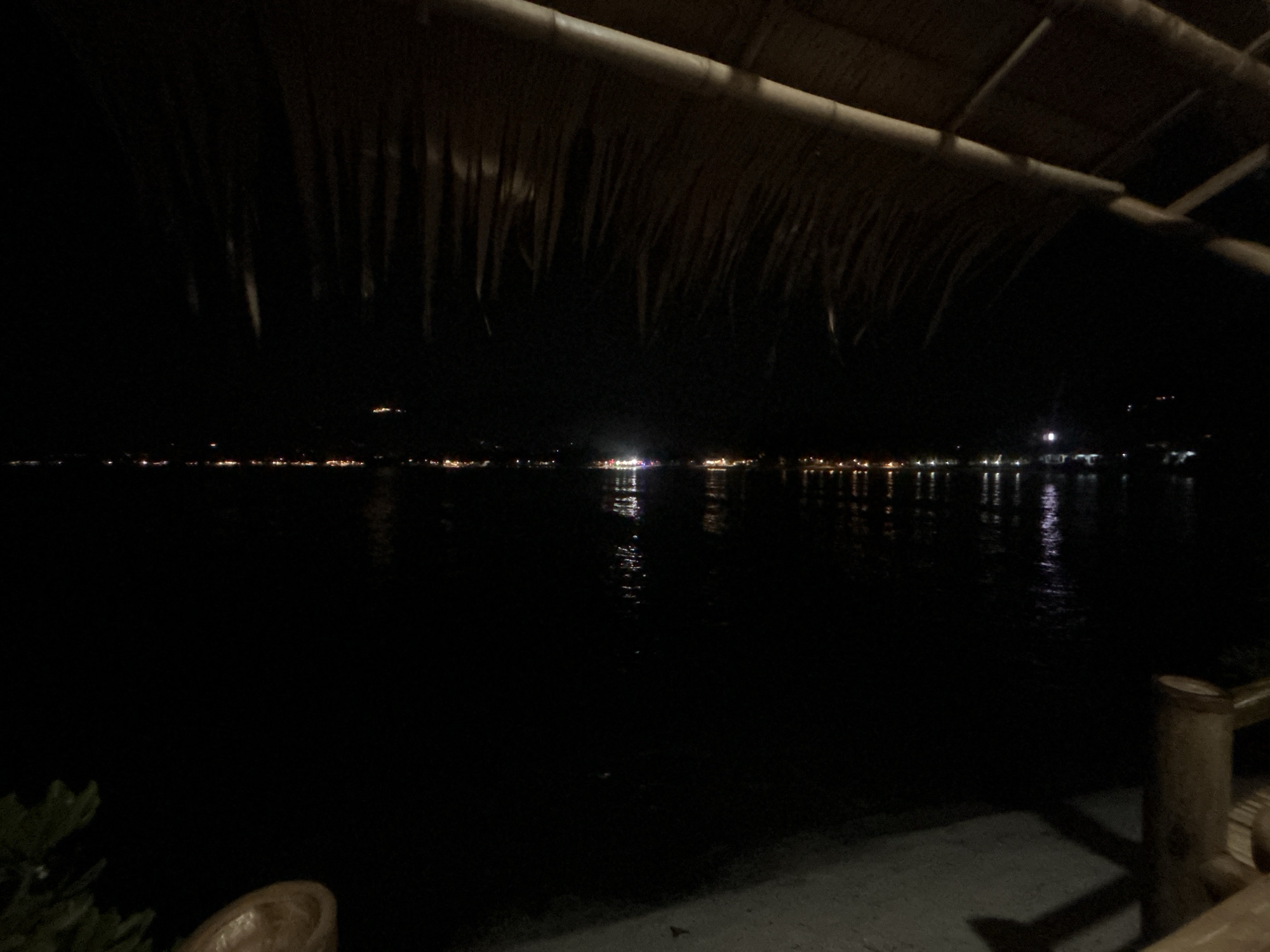 The image depicts a night scene from Baan Chaweng Noi, Thailand, likely near the coast. A thatched roof overhangs the view, partially obscuring the scene. In the distance, a line of lights indicates a settlement or shoreline. Reflections of these lights shimmer on the dark water. To the right, a wooden post and railing are visible. In the lower left, a small plant is present. The setting is dimly lit, with the primary source of light coming from the distant settlement. There are no people visible.