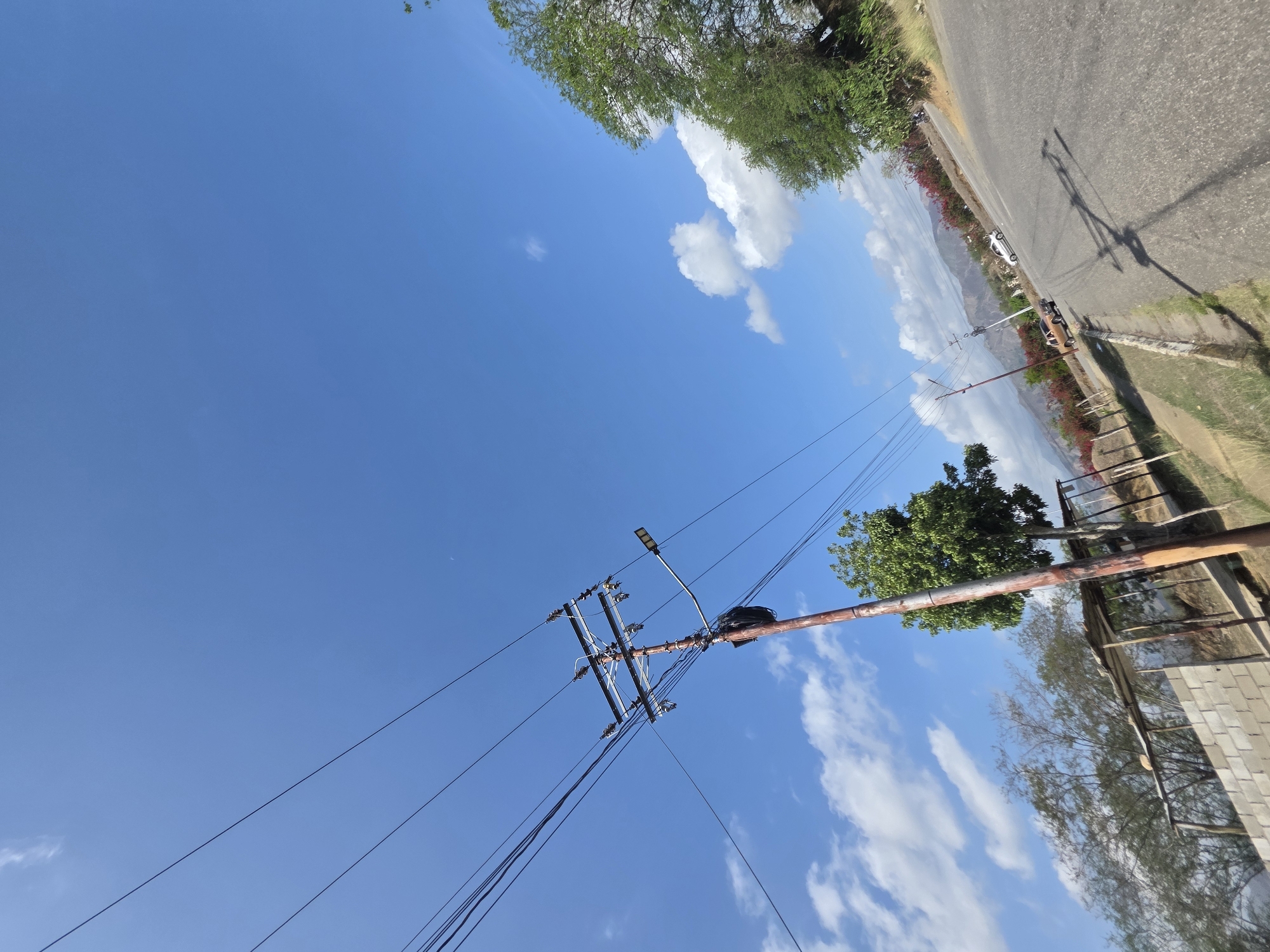 The image depicts a partly cloudy sky over Parroquia Ocumare, Venezuela. A utility pole with attached power lines is visible in the foreground. In the background, a road is lined with greenery. The camera angle suggests an upward view.