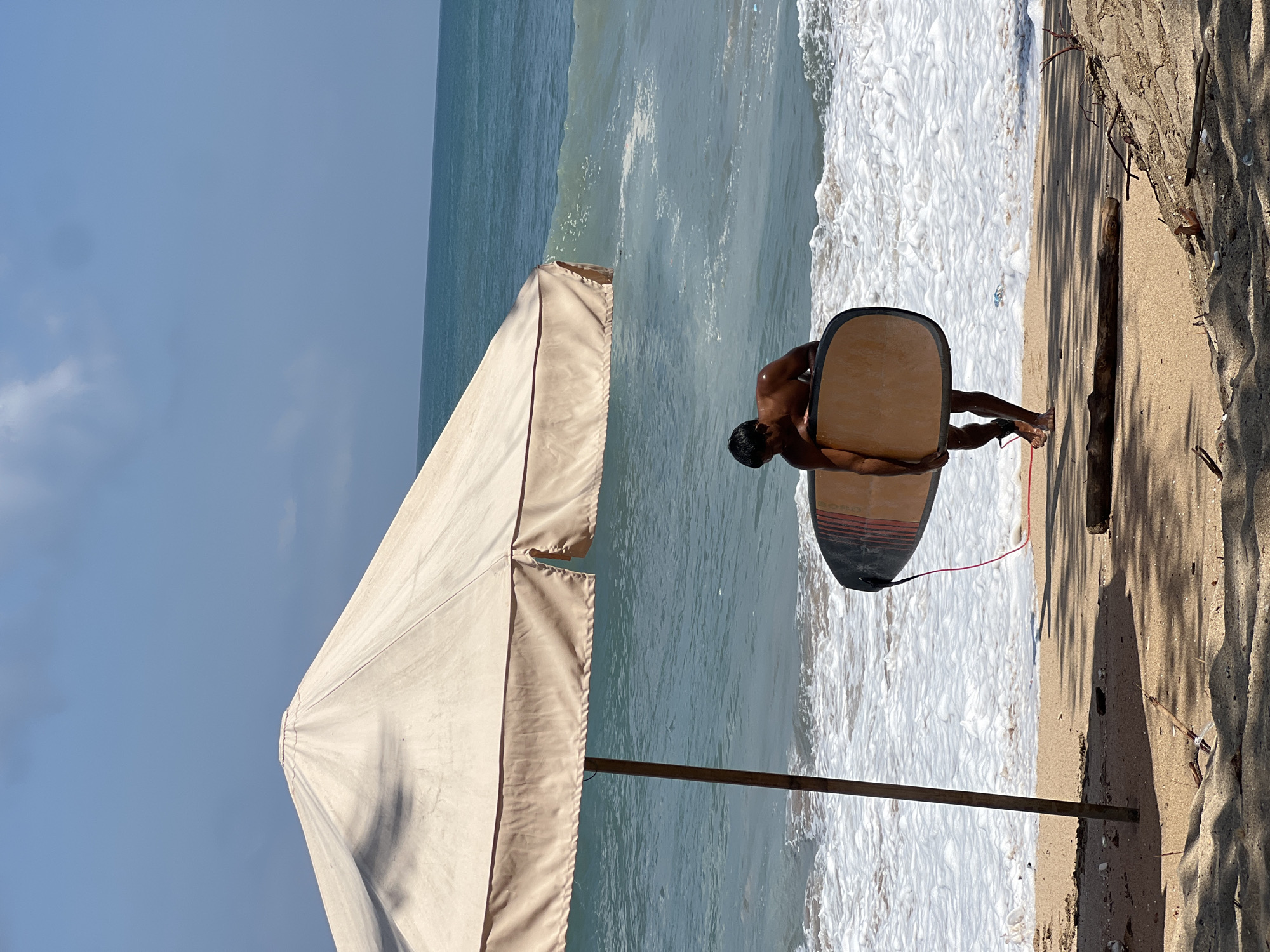 A shirtless person stands on a sandy beach in Kuta, Indonesia, holding a surfboard. The person is facing away from the camera and is partially obscured by a large, light-colored beach umbrella. The umbrella's pole is visible in the foreground. The ocean is in the background with waves breaking on the shore. The sky is blue with some clouds. There is a piece of driftwood on the sand in the middle ground. The lighting suggests it is daytime.
